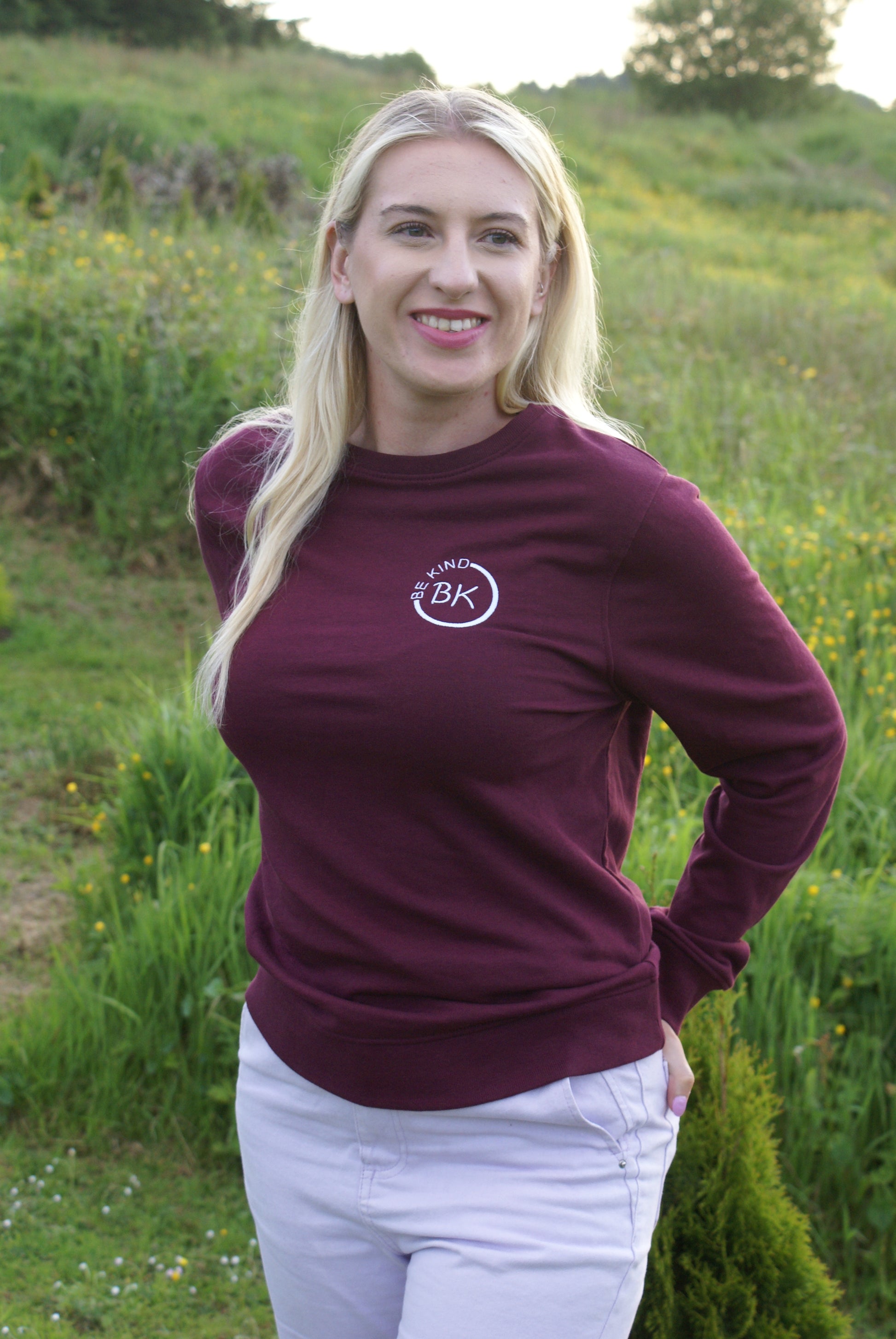 A woman stands in a field,  she's wearing an Organic Cotton Burgundy sweatshirt from Be Kind Apparel's Freestyle Range