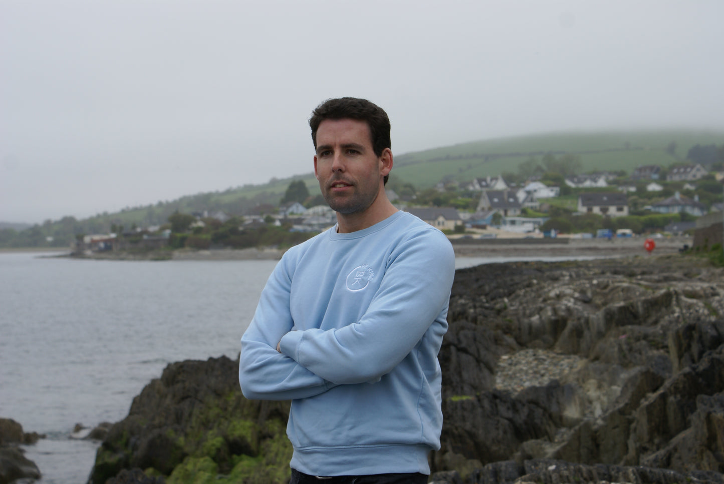 A man stands by the water, he's wearing a Sky Blue Organic Cotton Sweatshirt from the Be Kind Apparel Original Collection