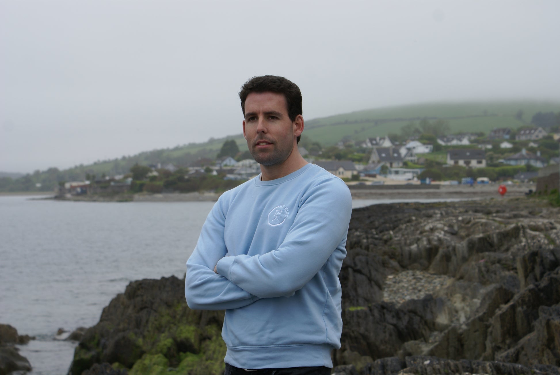A man stands by the water,  he's wearing a Sky Blue Organic Cotton Sweatshirt from the Be Kind Apparel Original Collection
