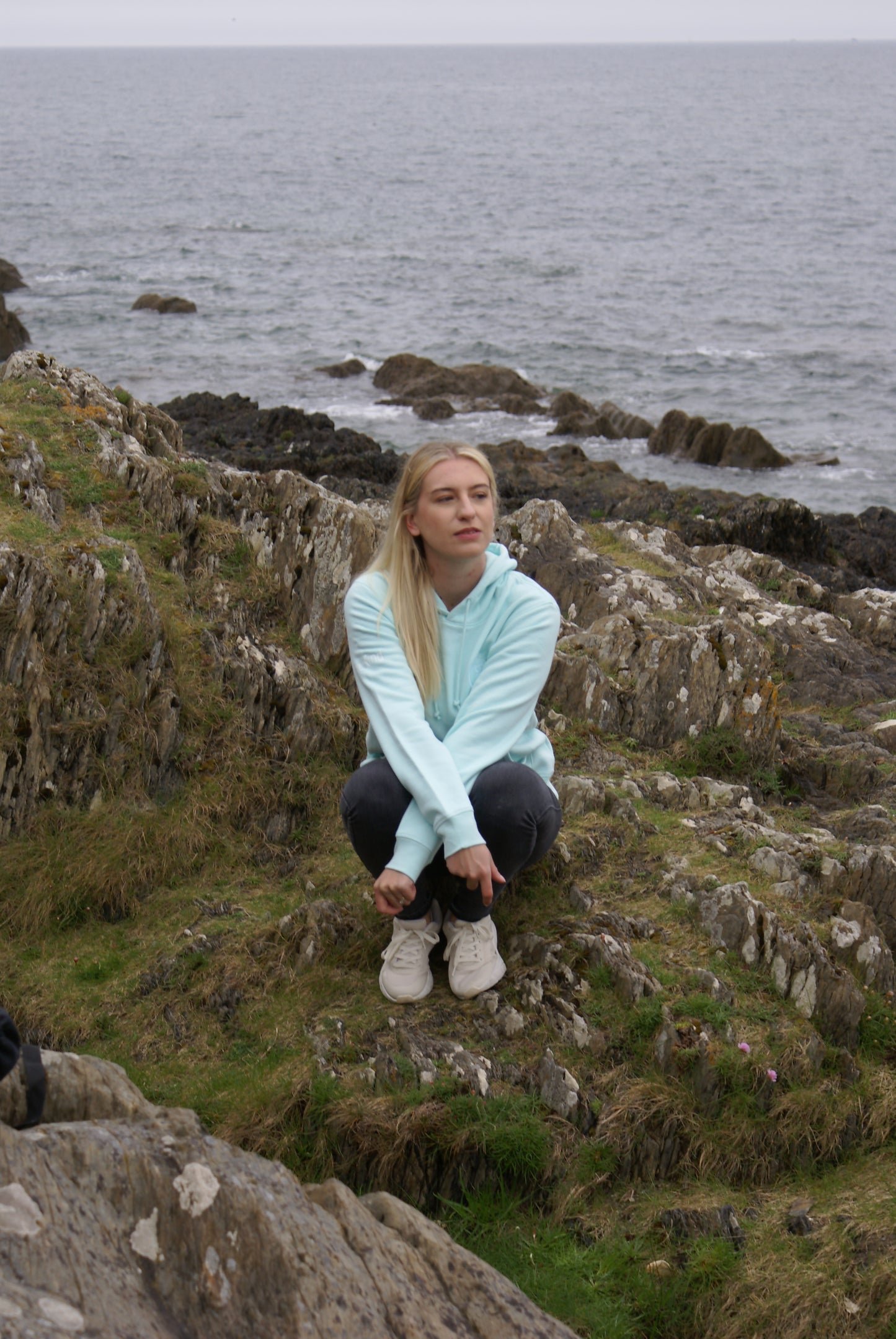 A woman sits on the shore in thought, she's wearing a Mint Green Organic Cotton Hoodie from the Be Kind Apparel Original Collection