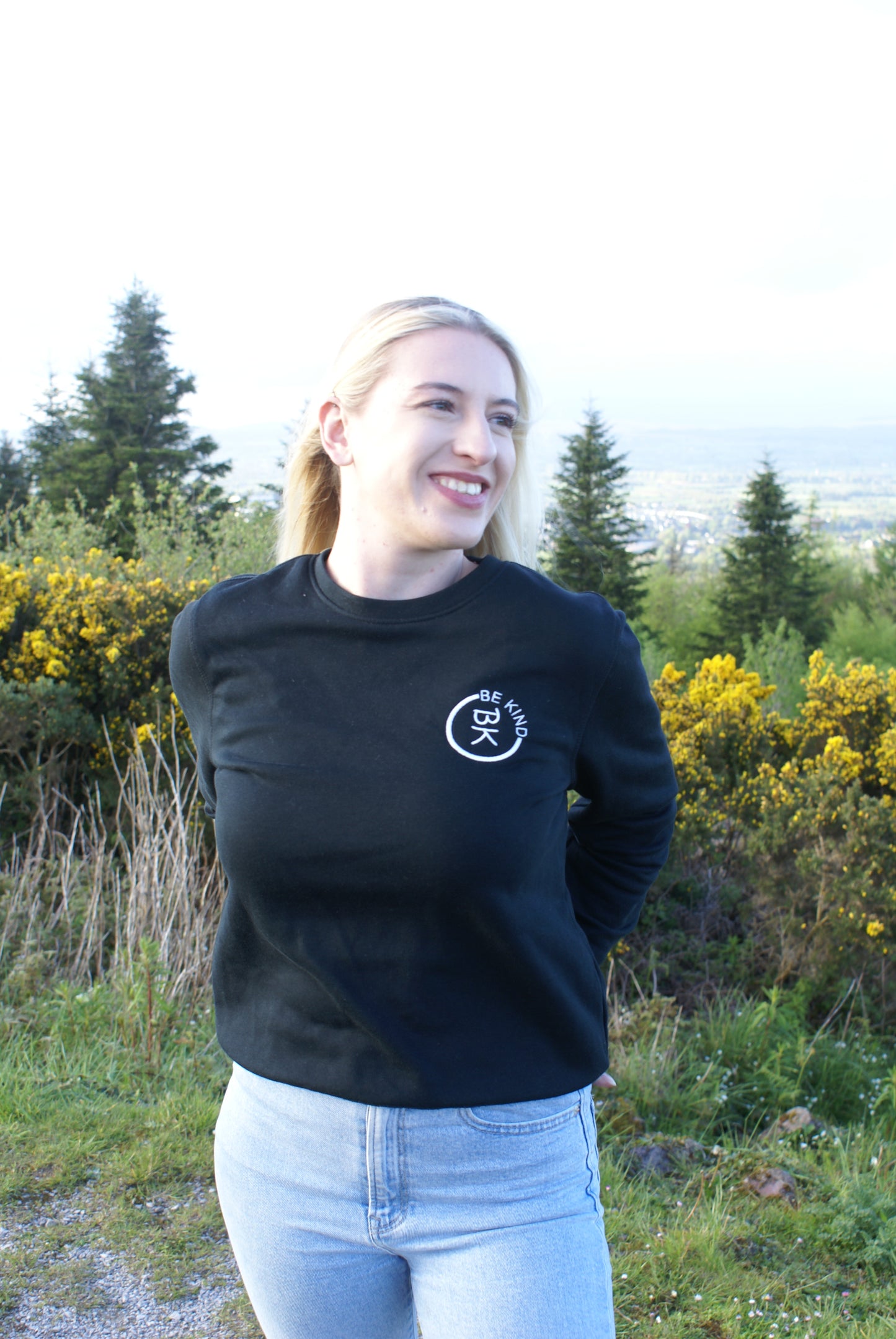 A woman has her hands behind her back, she's wearing a Black Organic Cotton Sweatshirt from the Be Kind Apparel Original Collection