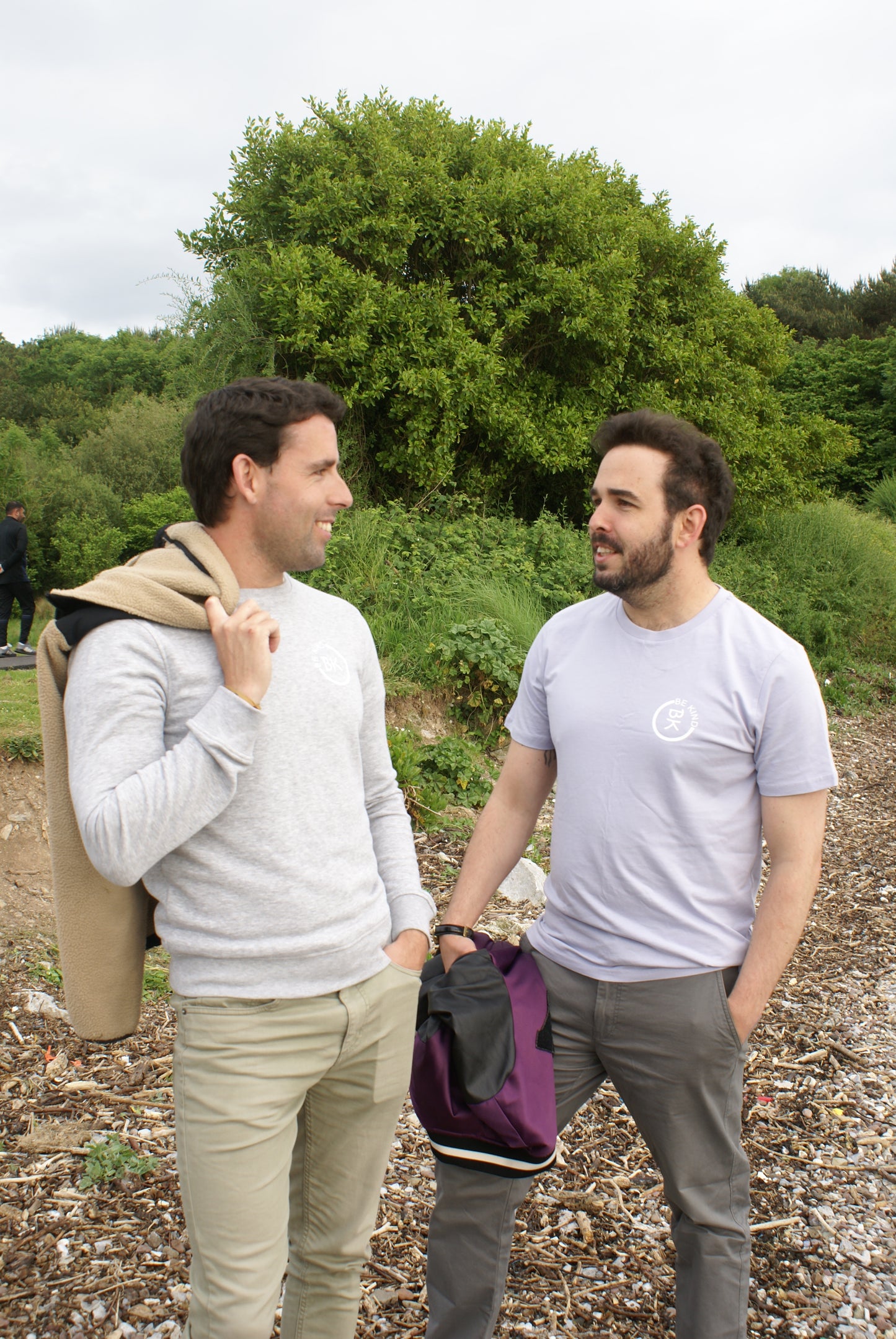 A man chats with his partner on a beach, he's wearing a Lavender Organic Cotton T-Shirt from the Be Kind Apparel Original Collection