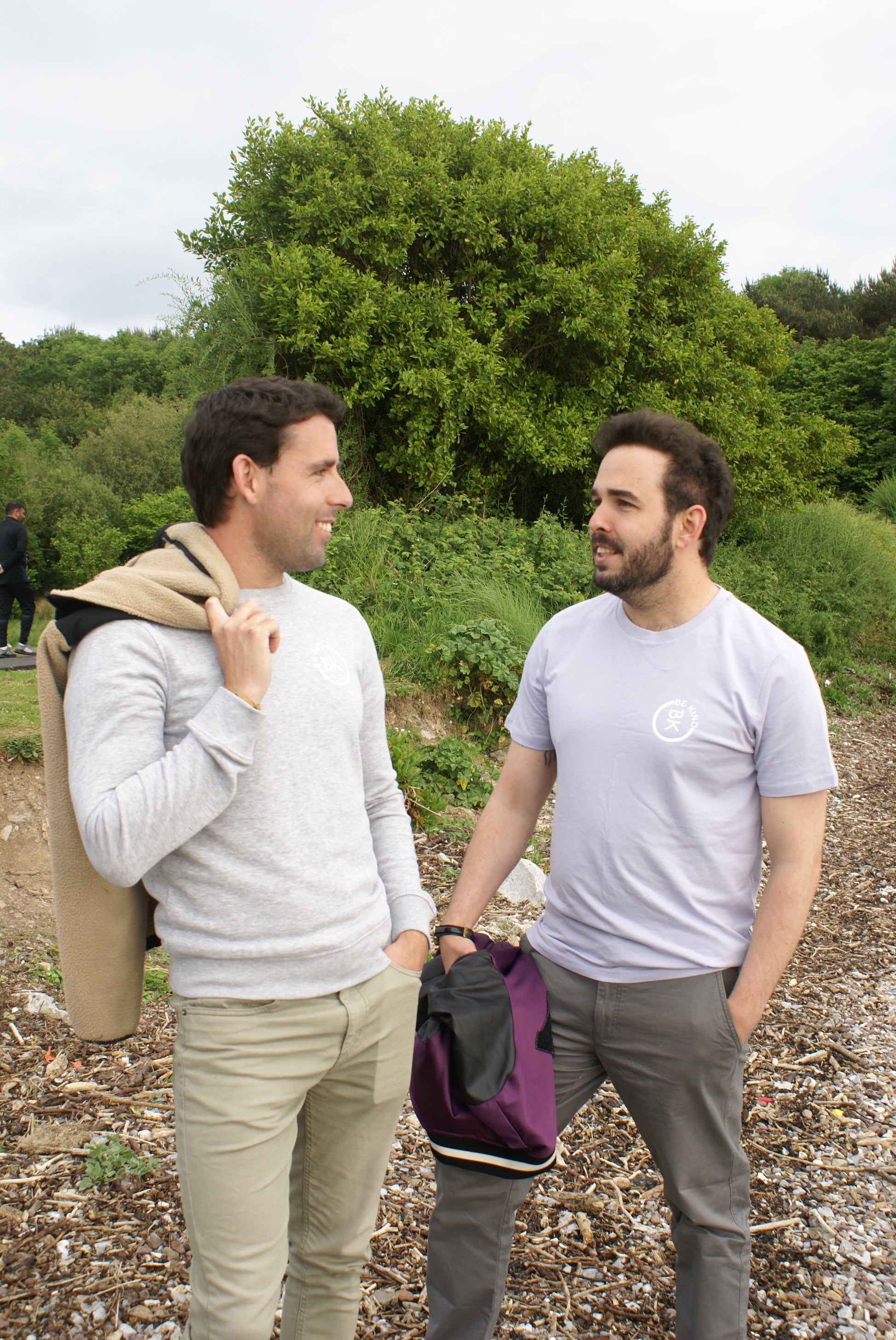 A man chats with his partner on a beach, he's wearing a Lavender Organic Cotton T-Shirt from the Be Kind Apparel Original Collection