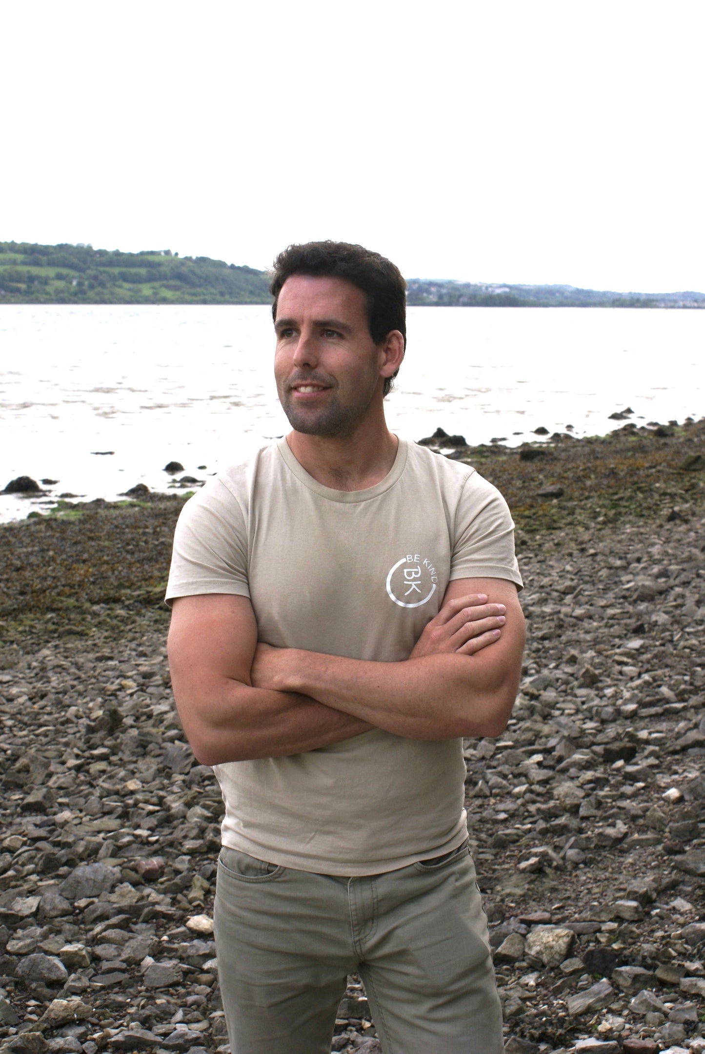A man stands on a beach, he's wearing a Sandstone Beige Organic Cotton T-Shirt from the Be Kind Apparel Original Collection