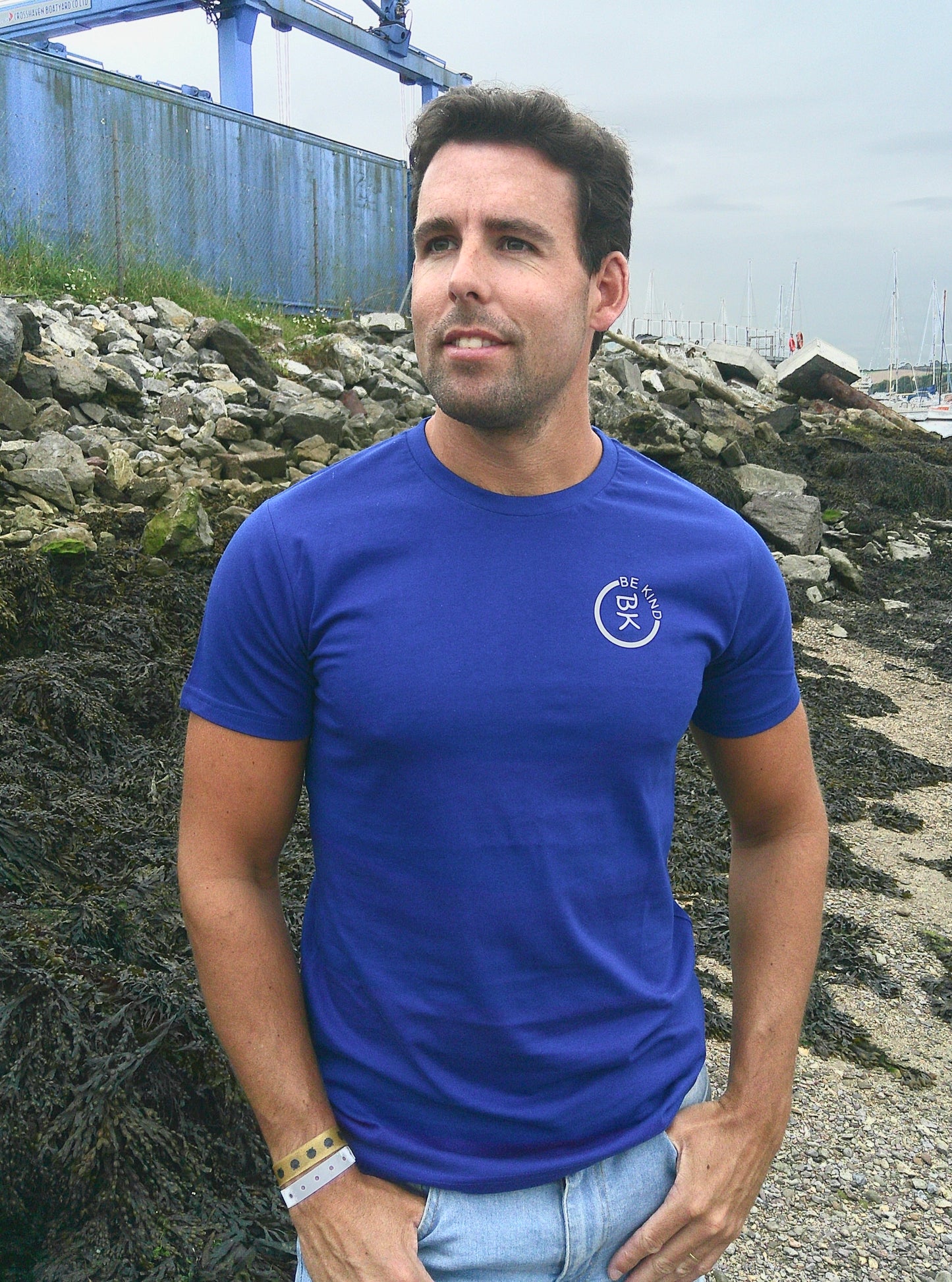 A man stands on the shore, he's wearing a Cobalt Blue Organic Cotton Connector T-Shirt from the Be Kind Apparel Connector Range