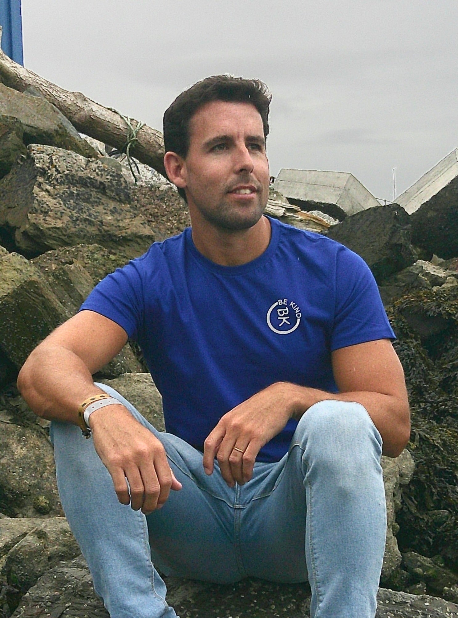 A man sits on a rock, he's wearing a Cobalt Blue Organic Cotton Connector T-Shirt from the Be Kind Apparel Connector Range