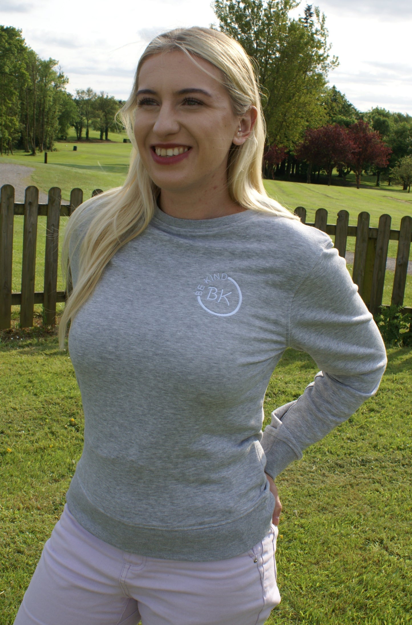 A woman stands by a fence, she's wearing a Grey Organic Cotton sweatshirt from Be Kind Apparel's Freestyle Range