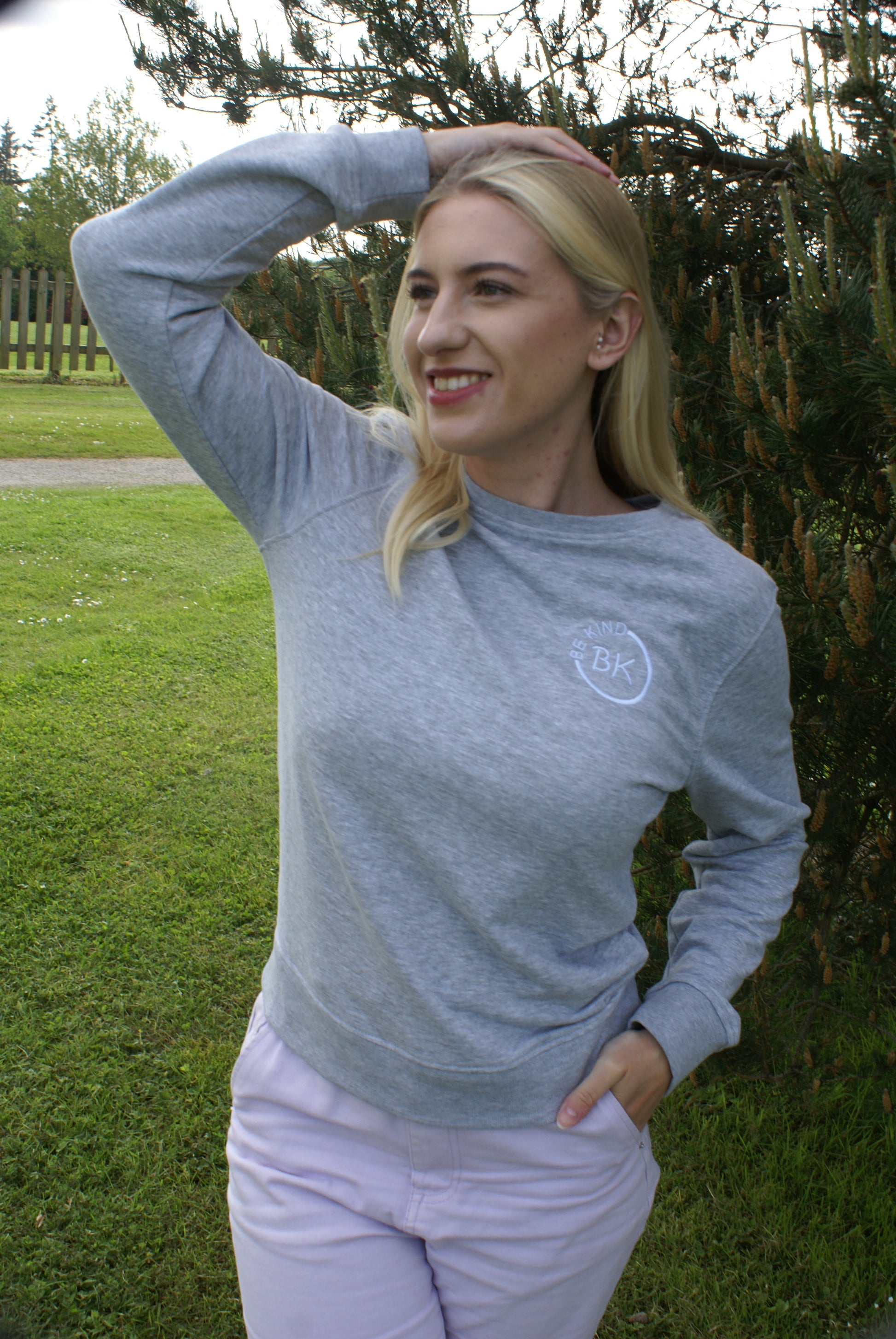A woman stands by a tree, she's wearing a Grey Organic Cotton sweatshirt from Be Kind Apparel's Freestyle Range