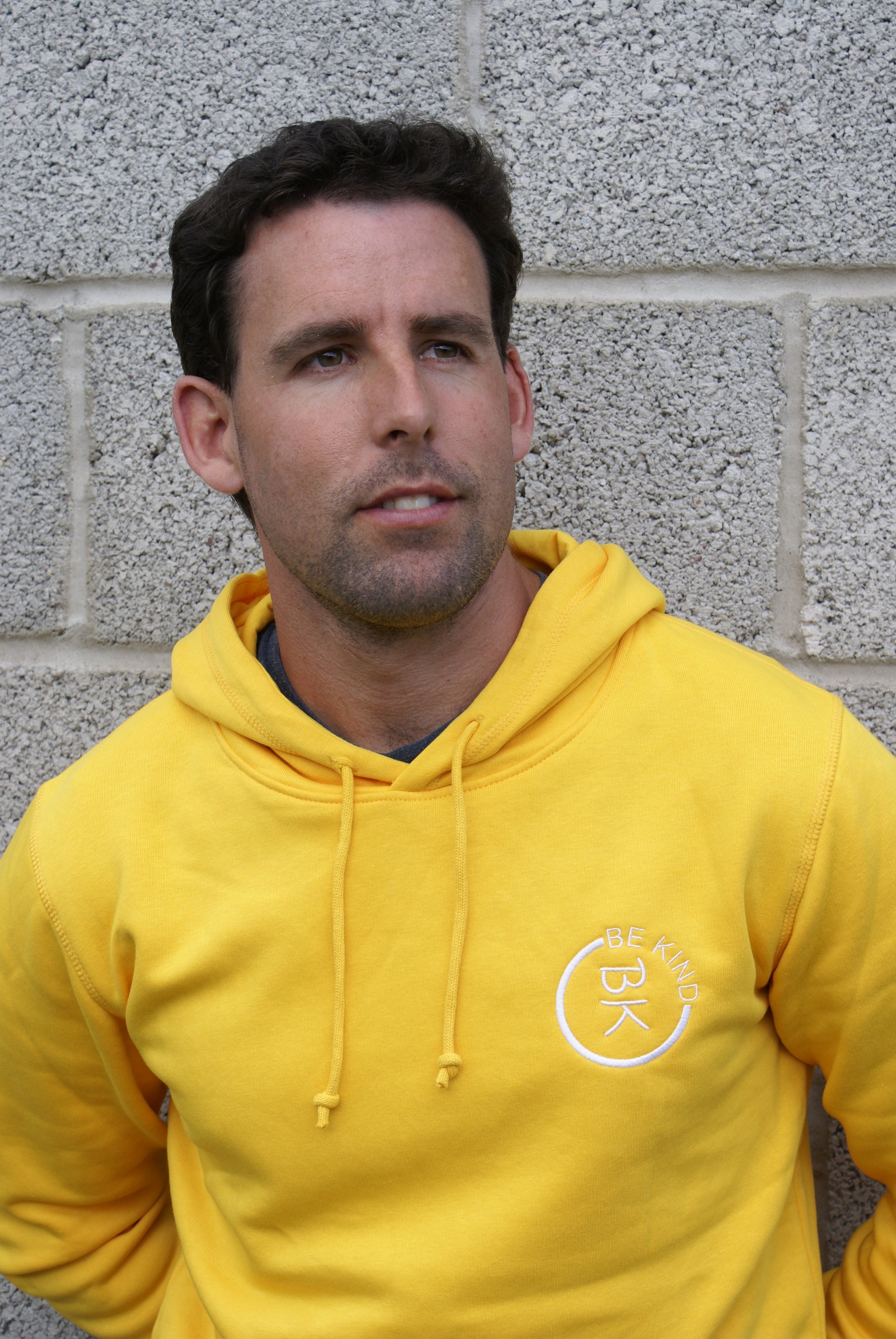A man leans against a wall, he's wearing a Sunshine Yellow Organic Cotton Hoodie from the Be Kind Apparel Original Collection