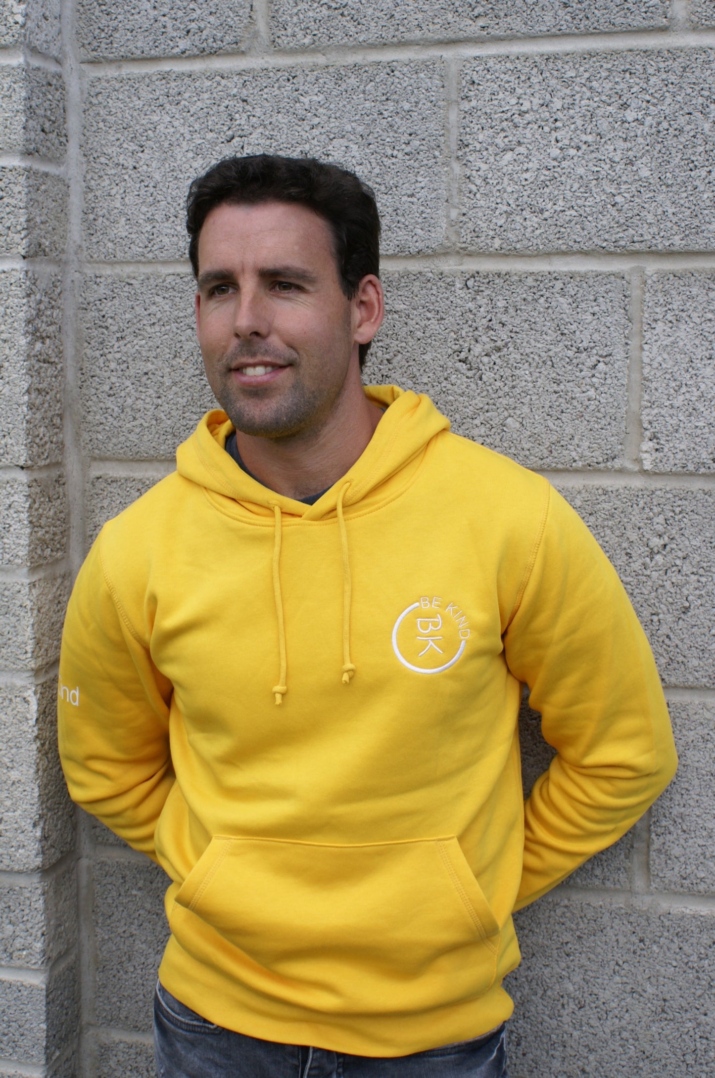 A man stands by a wall, he's wearing a Sunshine Yellow Organic Cotton Hoodie from the Be Kind Apparel Original Collection
