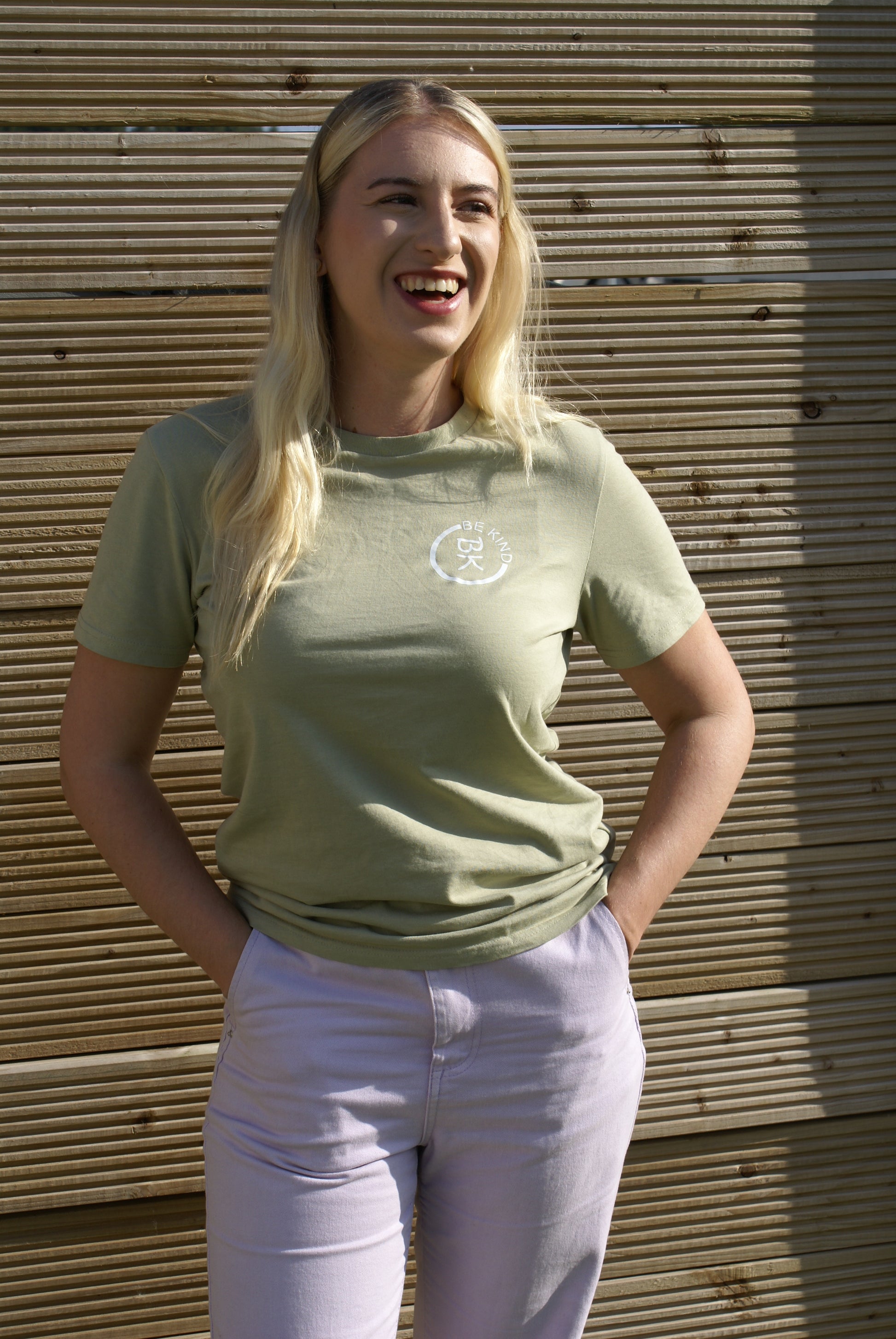 A woman stands laughing by a fence, she's wearing an Olive Green Organic Cotton T-Shirt from the Be Kind Apparel Original Collection