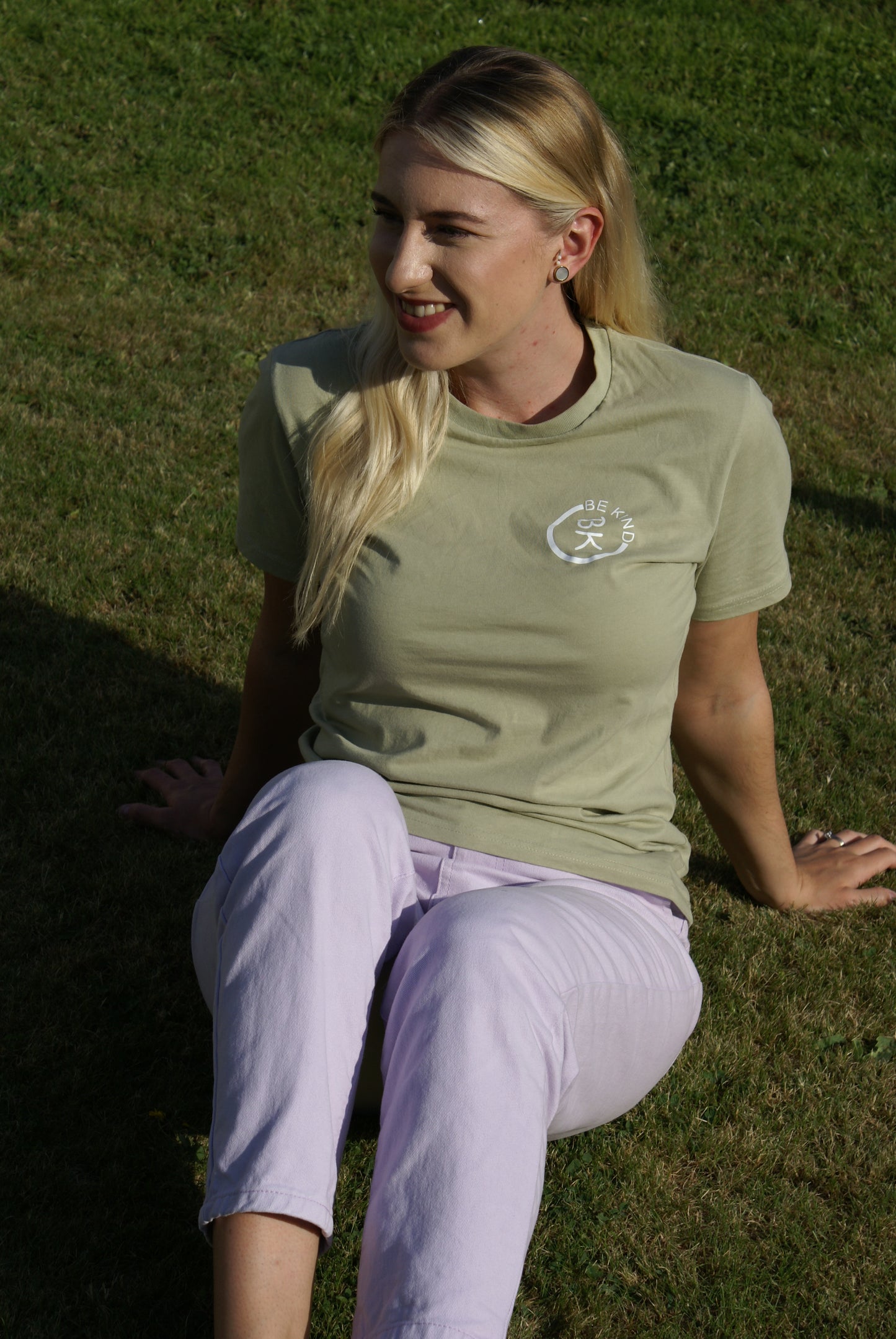 A woman sits in the grass, she's wearing an Olive Green Organic Cotton T-Shirt from the Be Kind Apparel Original Collection