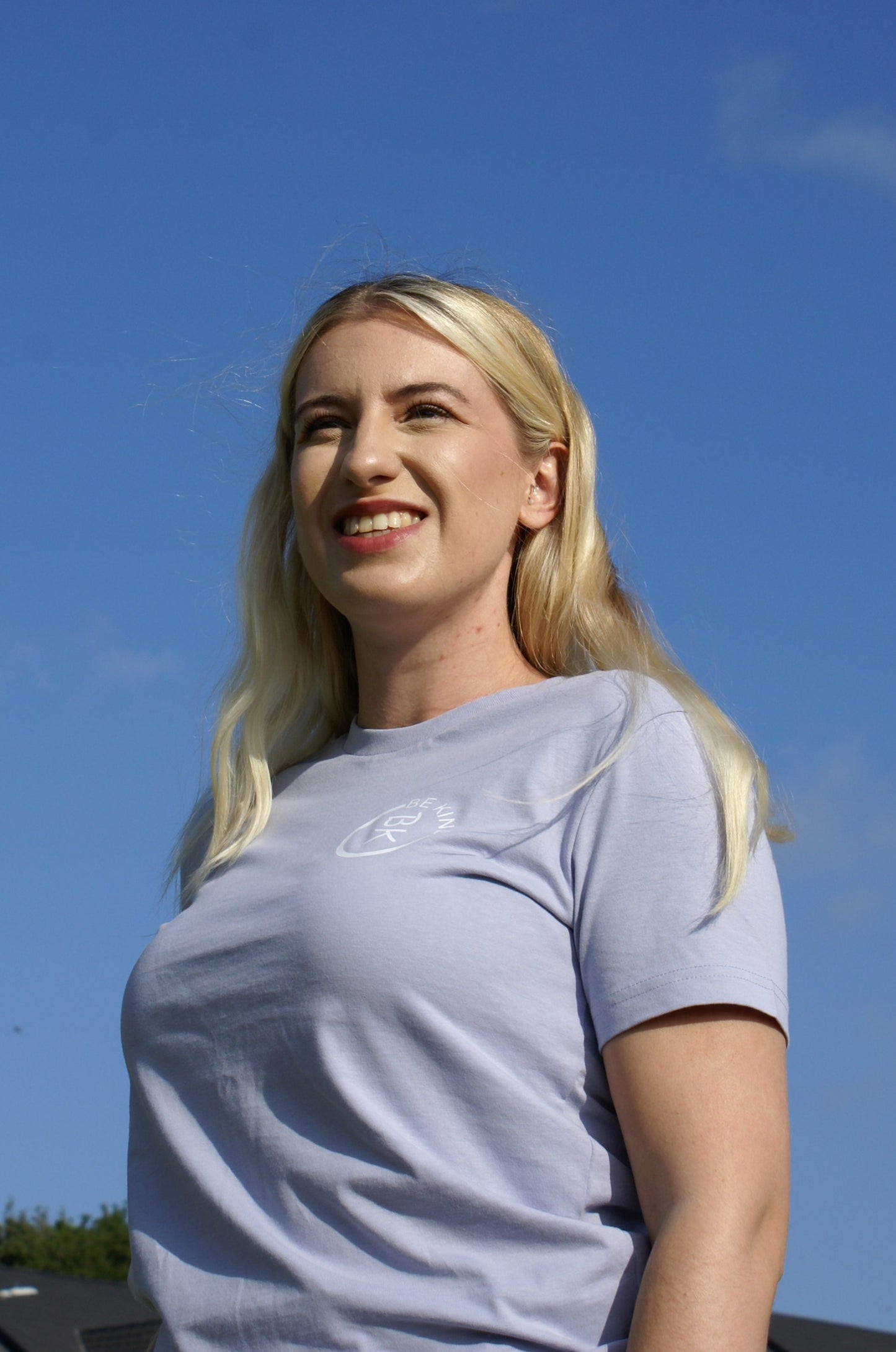 A woman smiles in the sun, she's wearing a Lavender Organic Cotton T-Shirt from the Be Kind Apparel Original Collection