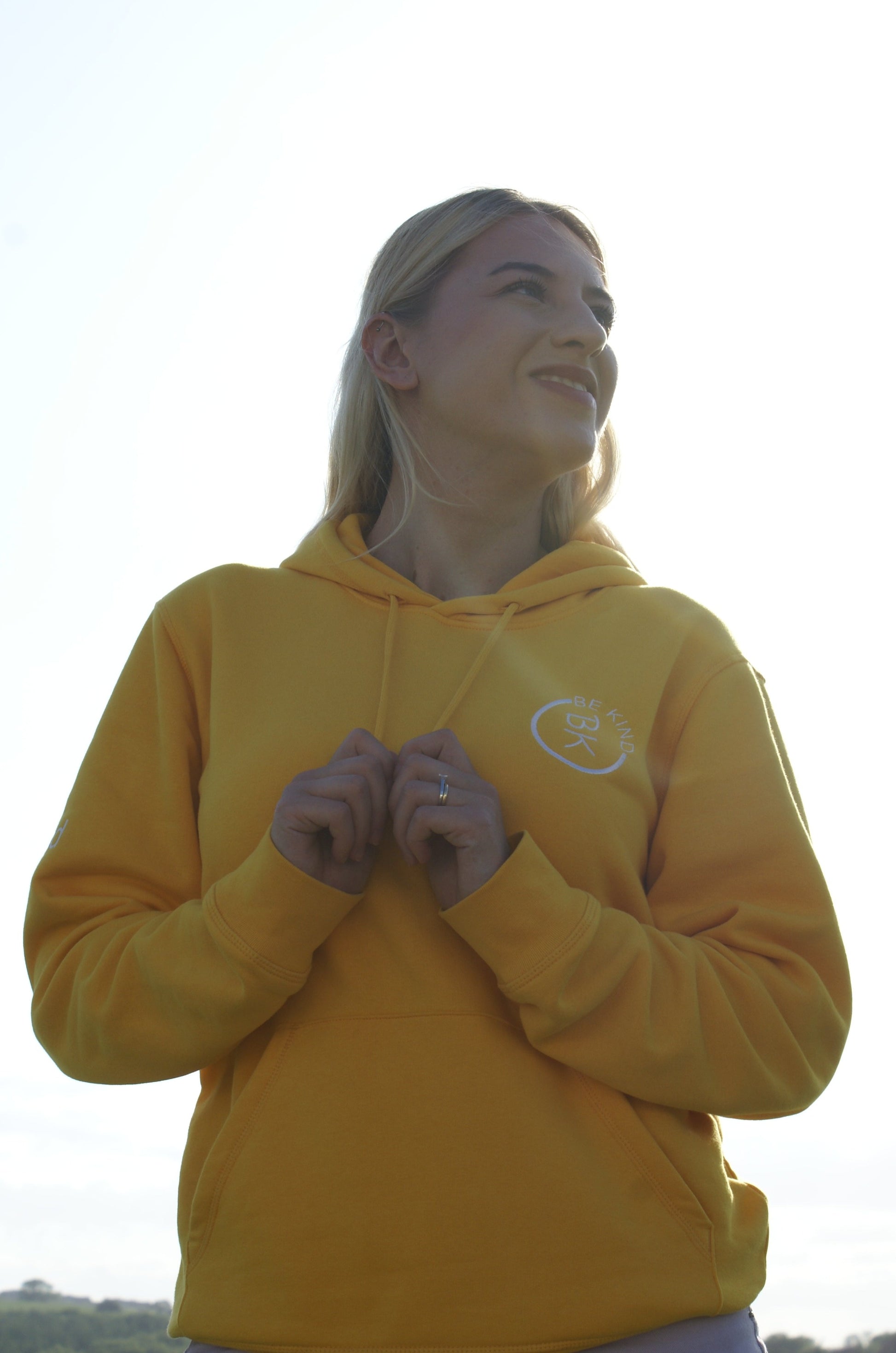 A woman stands, she's wearing a Sunshine Yellow Organic Cotton Hoodie from the Be Kind Apparel Original Collection