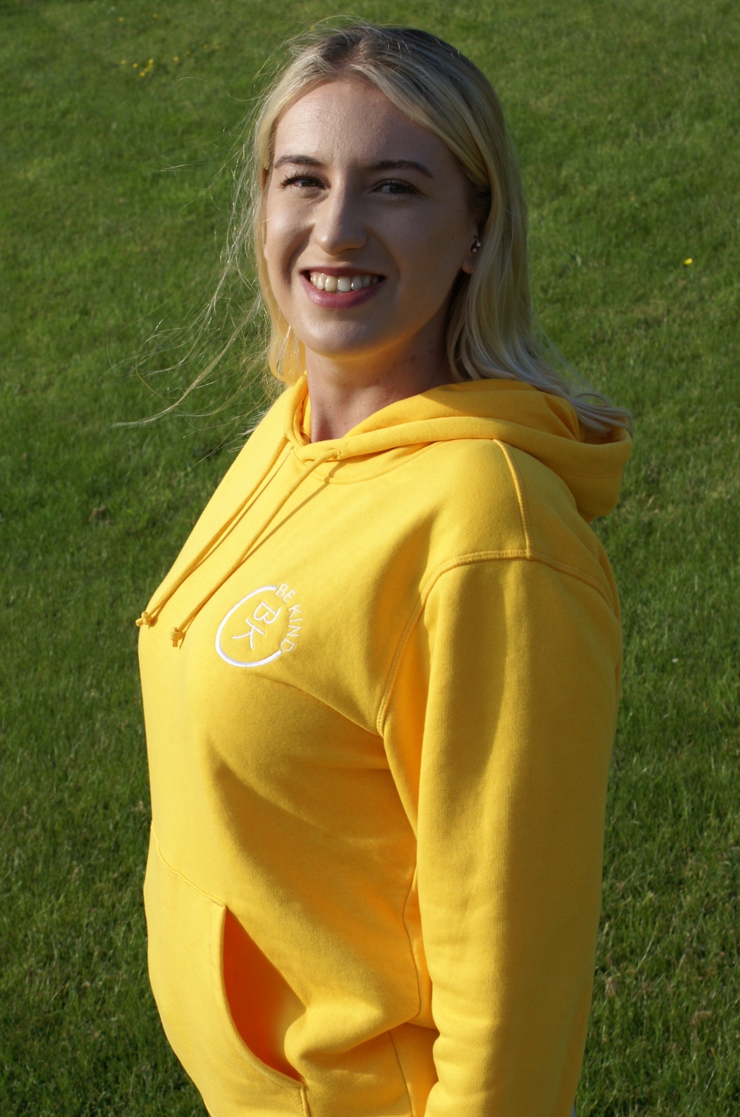 A woman smiles, she's wearing a Sunshine Yellow Organic Cotton Hoodie from the Be Kind Apparel Original Collection