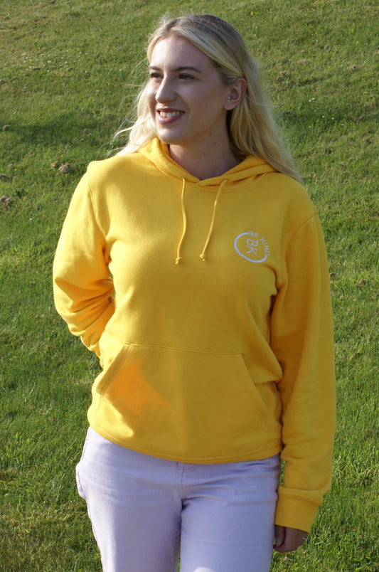 A woman stands in a field, she's wearing a Sunshine Yellow Organic Cotton Hoodie from the Be Kind Apparel Original Collection