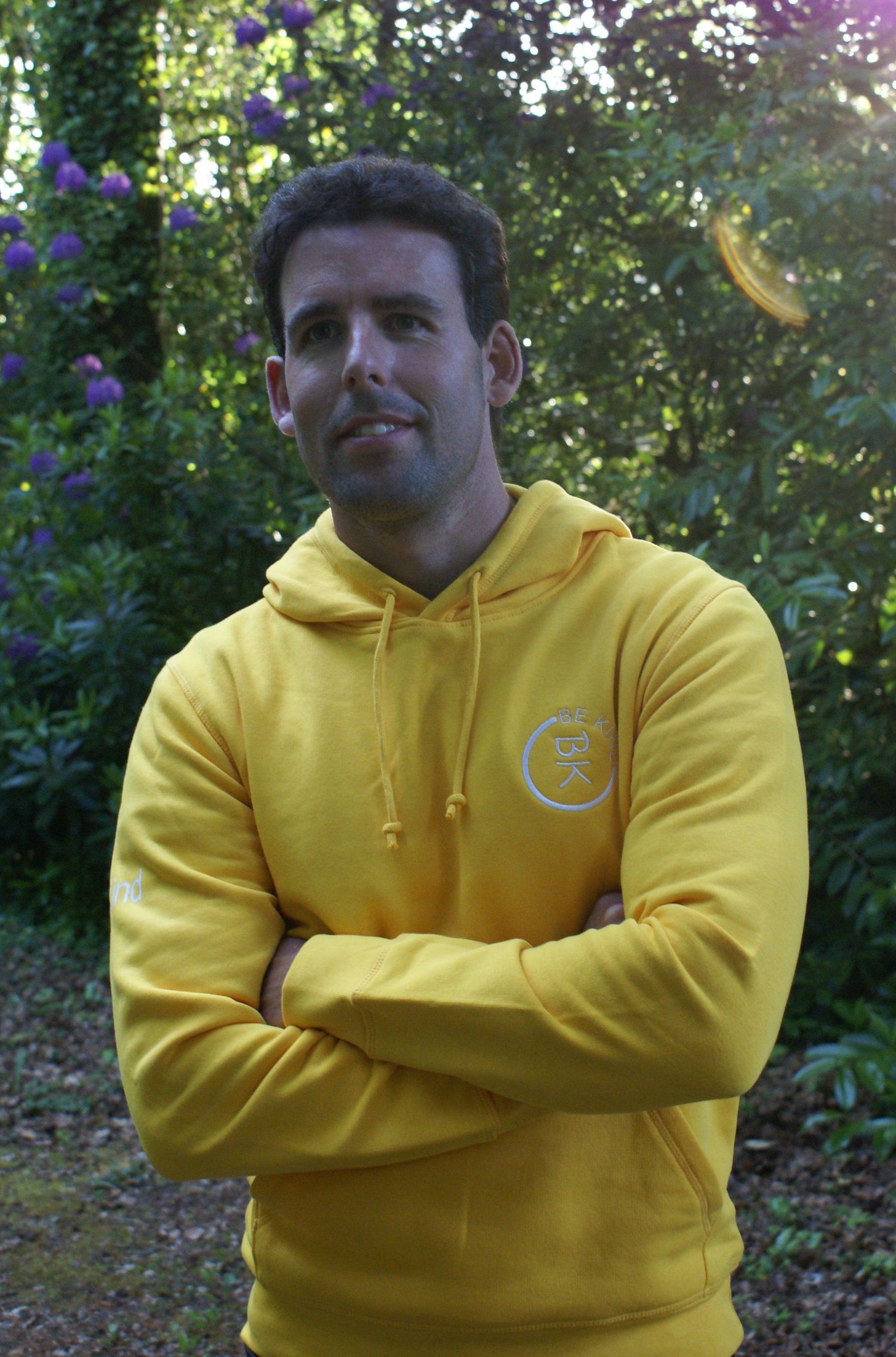 A man stands in a forest, he's wearing a Sunshine Yellow Organic Cotton Hoodie from the Be Kind Apparel Original Collection