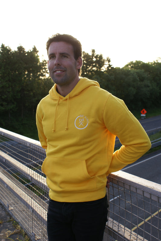 A man stands on a bridge, he's wearing a Sunshine Yellow Organic Cotton Hoodie from the Be Kind Apparel Original Collection