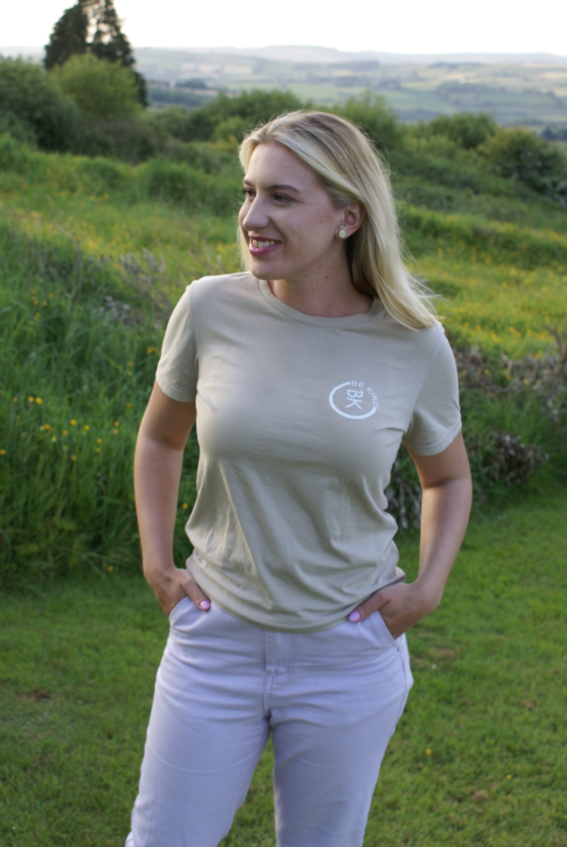 A woman stands with her hands in her pockets, she's wearing a Sandstone Beige Organic Cotton T-Shirt from the Be Kind Apparel Original Collection