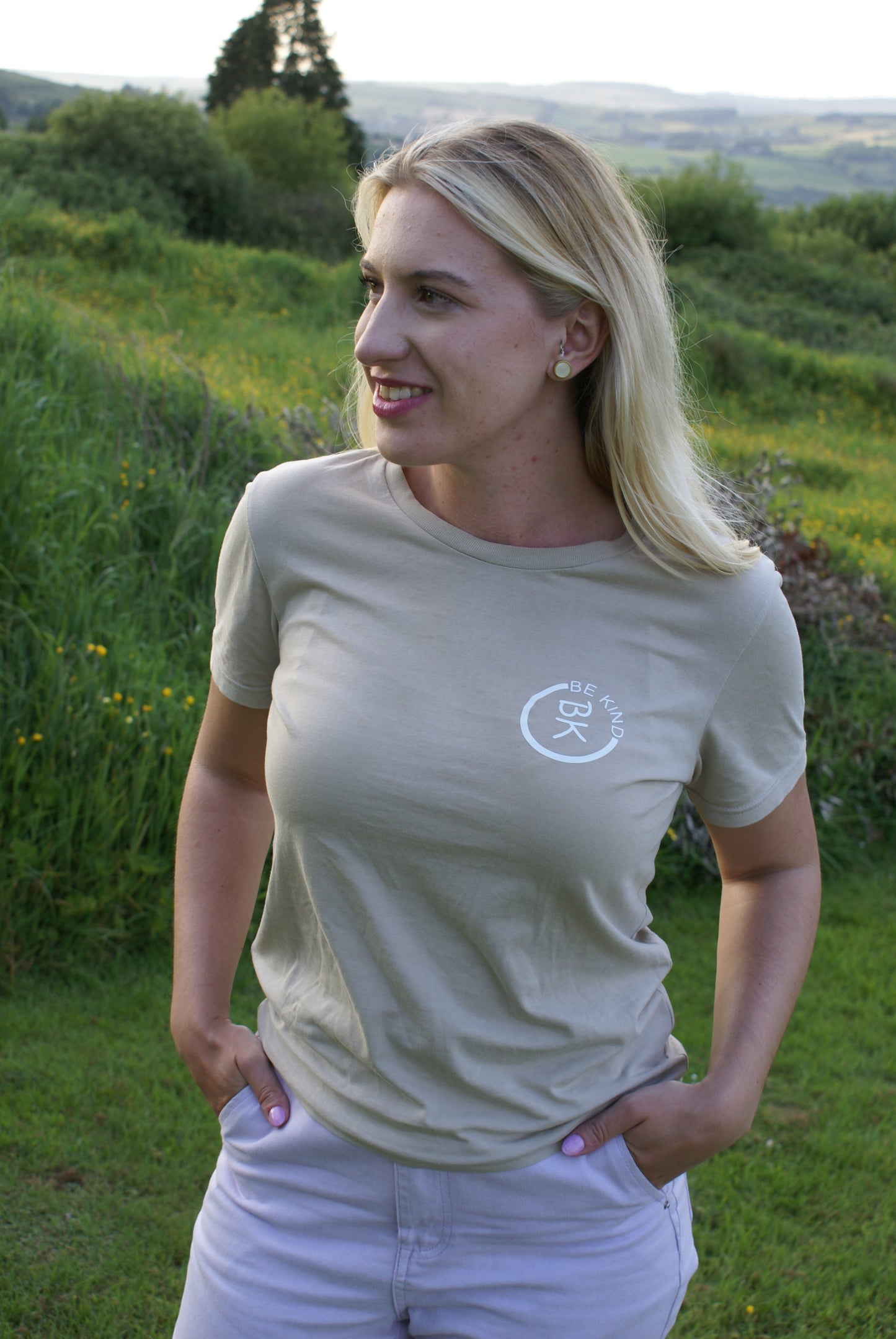 A woman stands in a green field, she's wearing a Sandstone Beige Organic Cotton T-Shirt from the Be Kind Apparel Original Collection