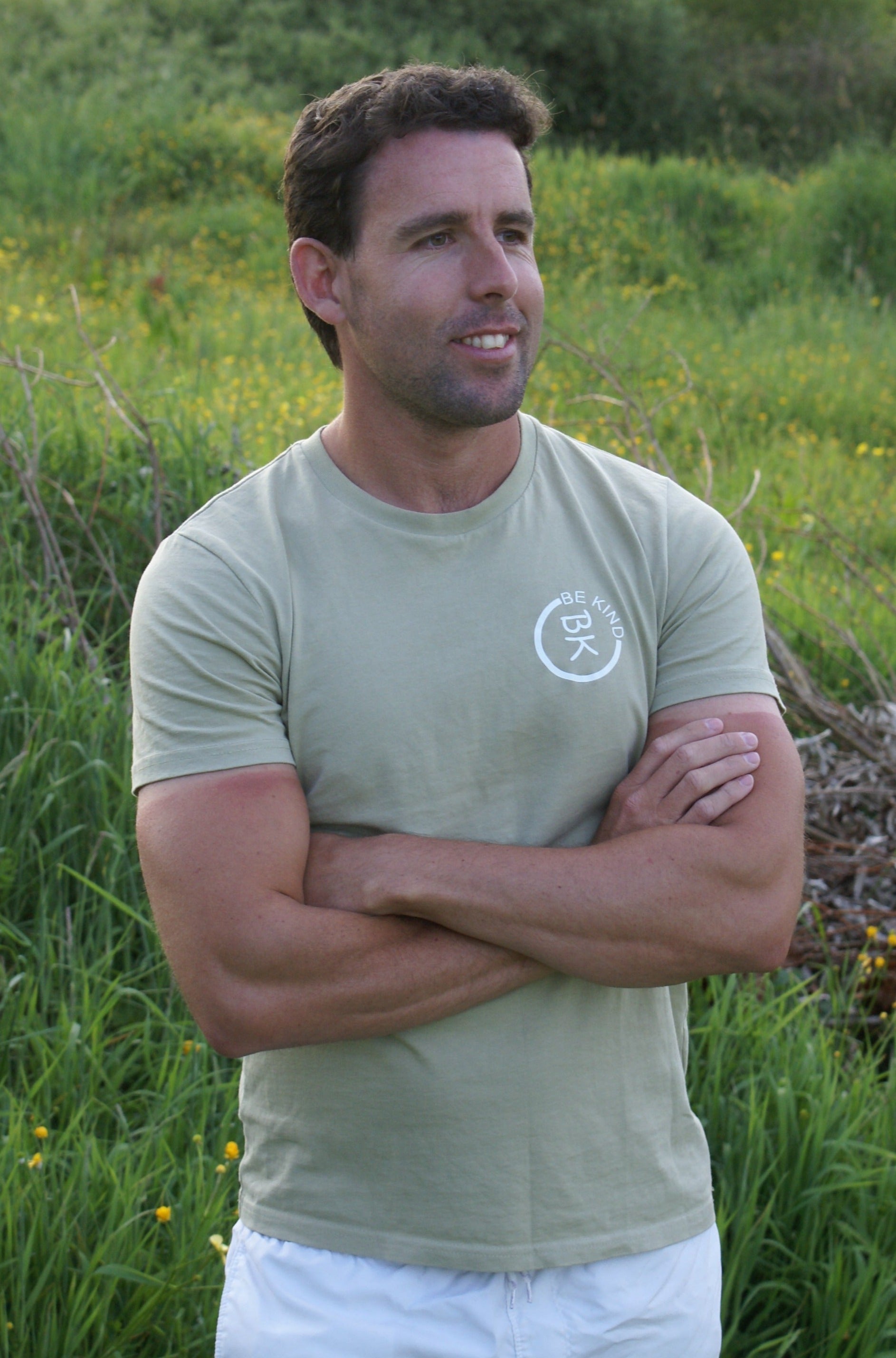 A man stands in a field, he's wearing an Olive Green Organic Cotton T-Shirt from the Be Kind Apparel Original Collection