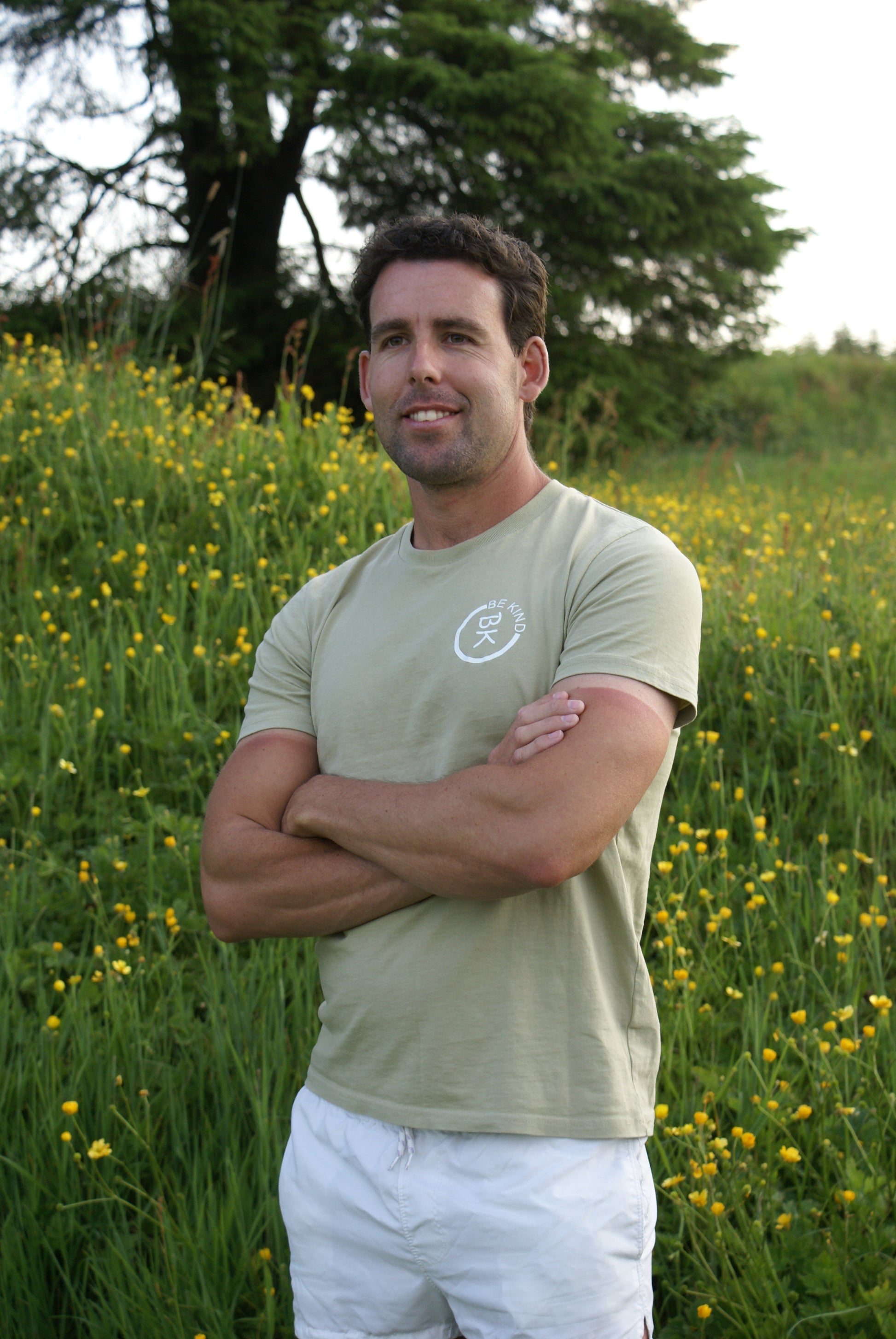 A man stands in a field of flowers, he's wearing an Olive Green Organic Cotton T-Shirt from the Be Kind Apparel Original Collection