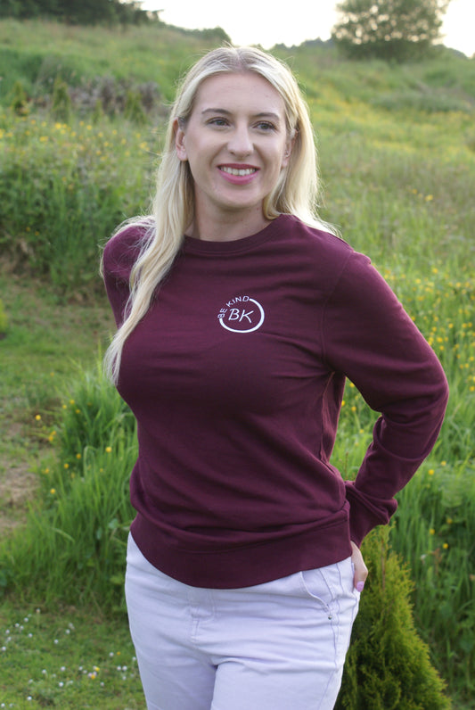 A woman stands in a field,  she's wearing an Organic Cotton Burgundy sweatshirt from Be Kind Apparel's Freestyle Range