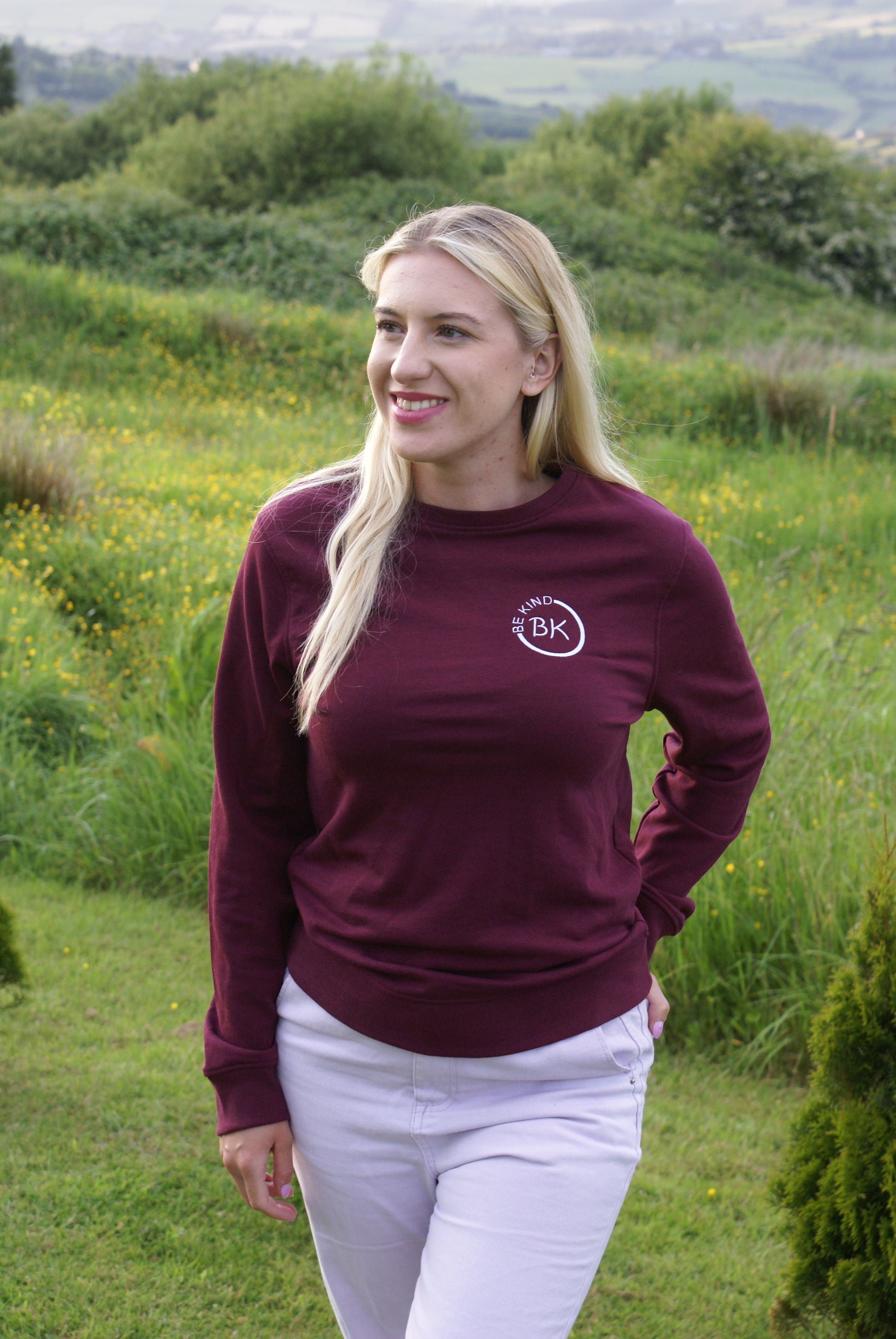 A woman stands in a field, she's wearing an Organic Cotton Burgundy sweatshirt from Be Kind Apparel's Freestyle Range