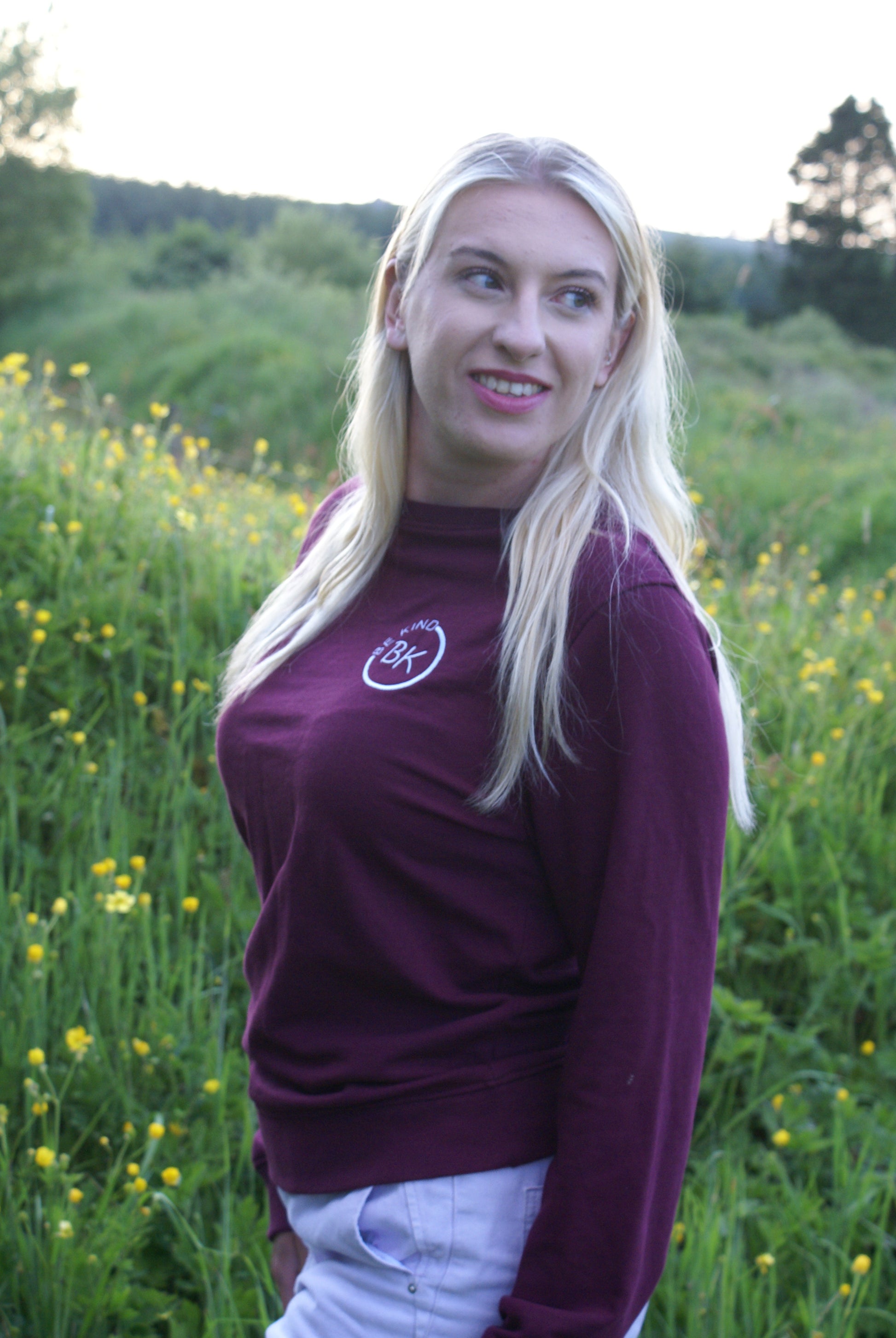 A woman looks back in a field full of flowers. she's wearing an Organic Cotton Burgundy sweatshirt from Be Kind Apparel's Freestyle Range