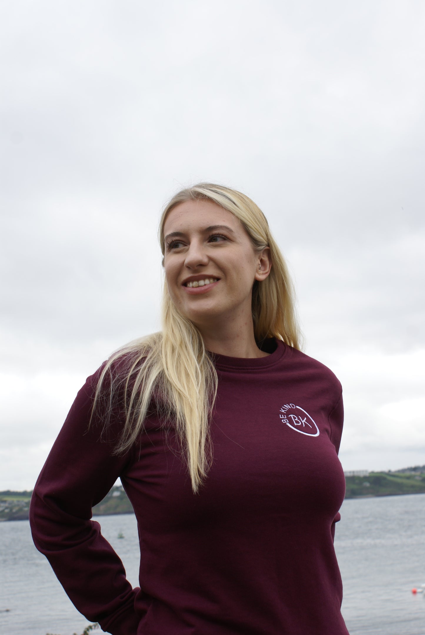 A woman stands by the ocean, she's wearing an Organic Cotton Burgundy sweatshirt from Be Kind Apparel's Freestyle Range