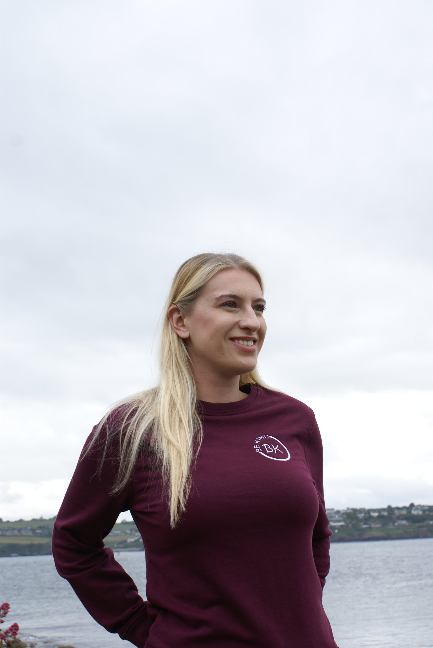 A woman stands by the water, she's wearing an Organic Cotton Burgundy sweatshirt from Be Kind Apparel's Freestyle Range