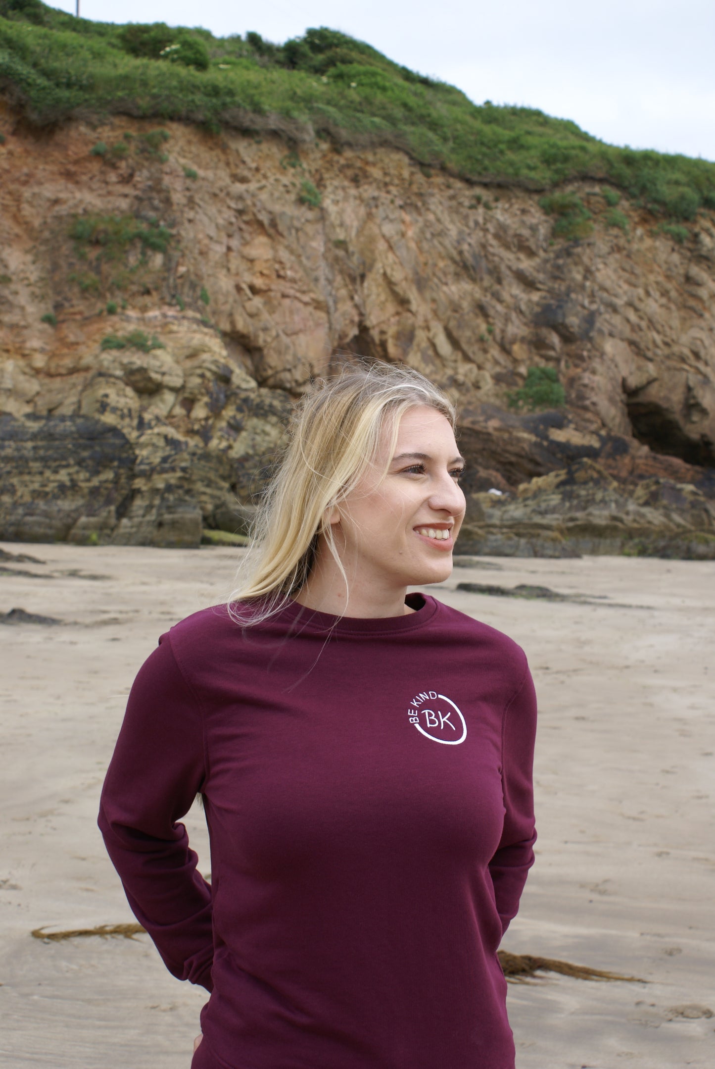 A woman stands on a beach, she's wearing an Organic Cotton Burgundy sweatshirt from Be Kind Apparel's Freestyle Range