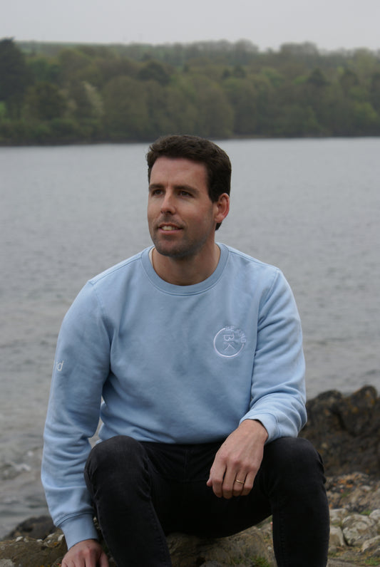 A man sits by the shore, he's wearing a Sky Blue Organic Cotton Sweatshirt from the Be Kind Apparel Original Collection