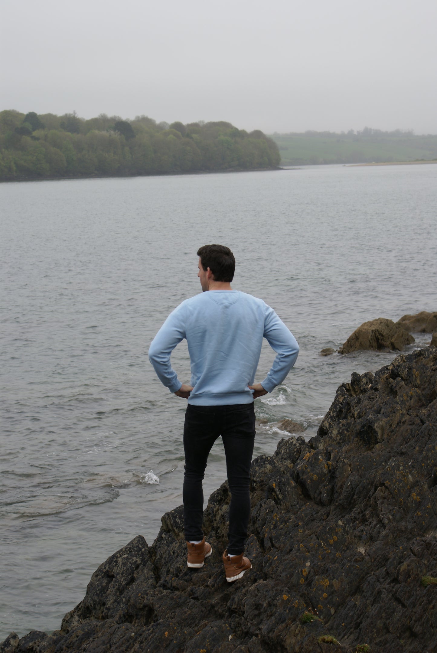 A man with hands on hips stands by a river,  he's wearing a Sky Blue Organic Cotton Sweatshirt from the Be Kind Apparel Original Collection
