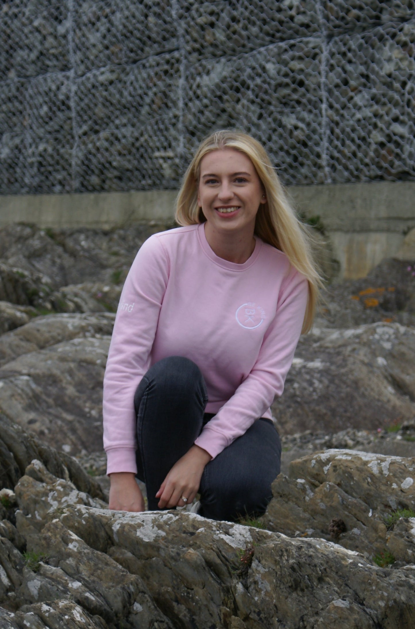 A woman kneels on some rocks, she's wearing a Bubblegum Pink Organic Cotton Sweatshirt from the Be Kind Apparel Original Collection