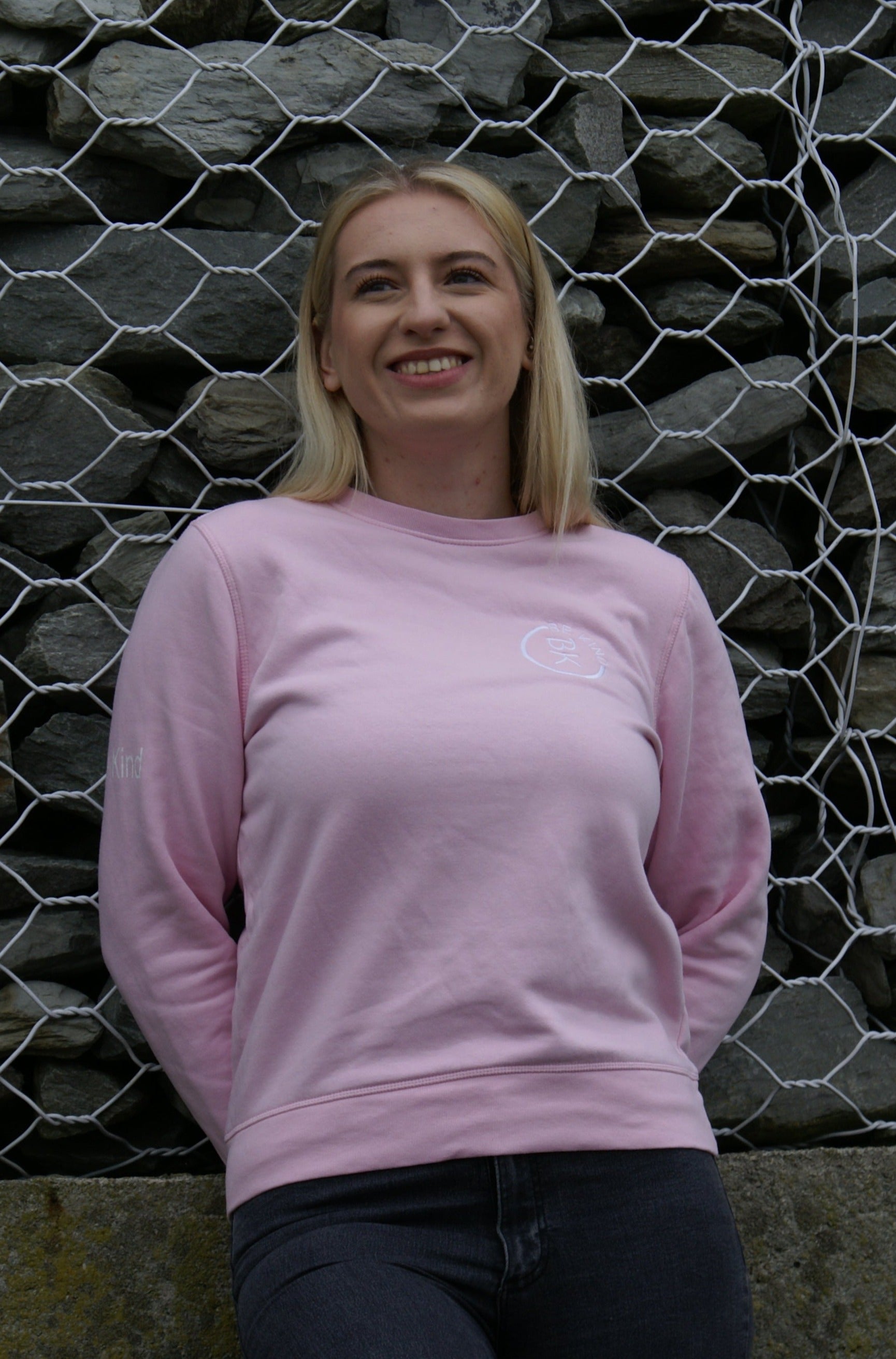A woman leans against a rock wall, she's wearing a Bubblegum Pink Organic Cotton Sweatshirt from the Be Kind Apparel Original Collection