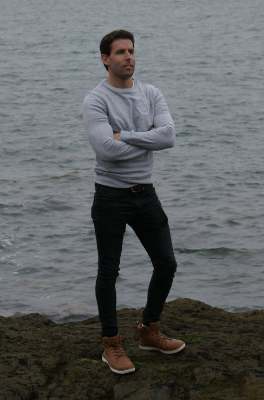 A man stands next to the water with his arms folded, he's wearing a Grey Organic Cotton sweatshirt from Be Kind Apparel's Freestyle Range