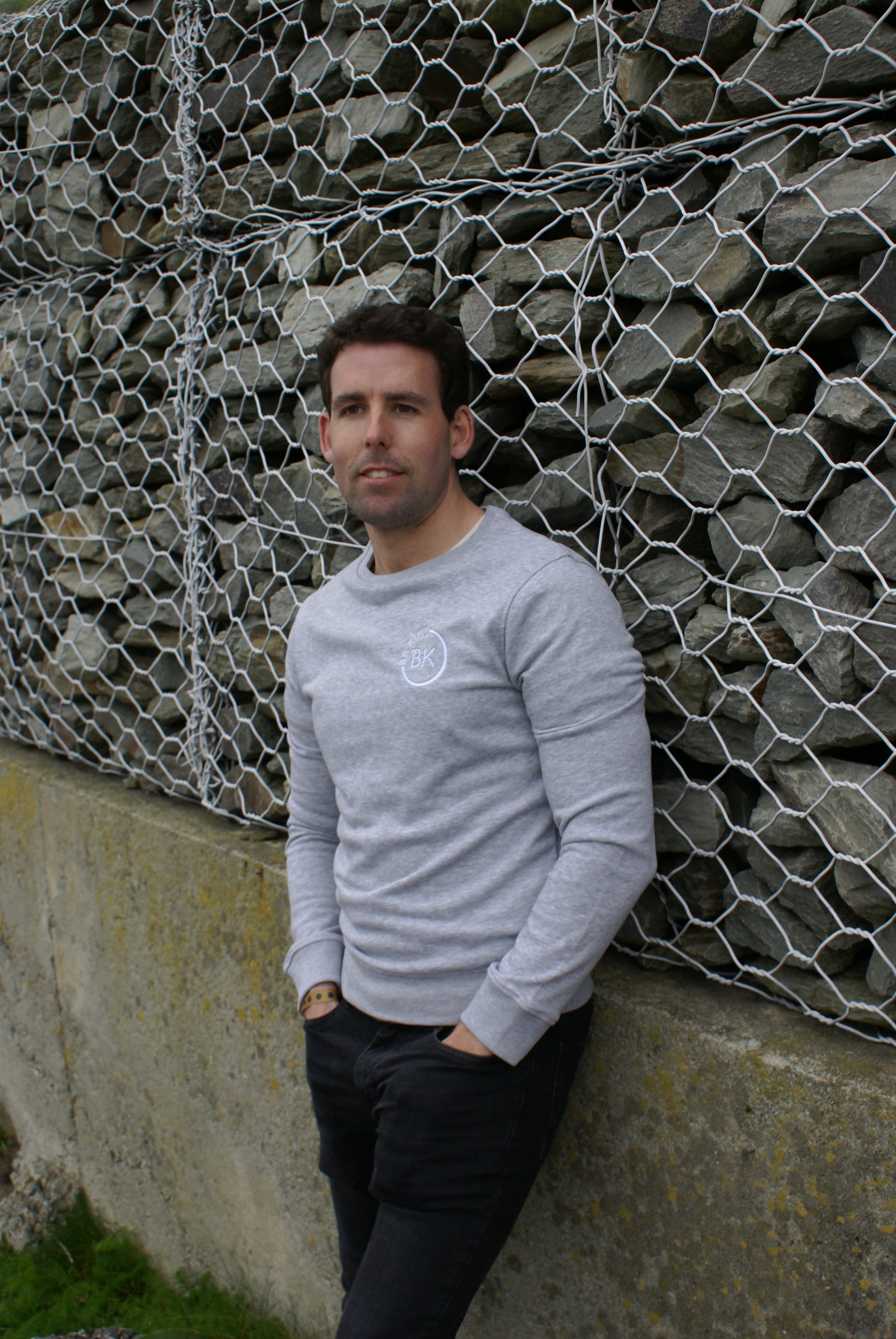 A man leans on a wall, he's wearing a Grey Organic Cotton sweatshirt from Be Kind Apparel's Freestyle Range