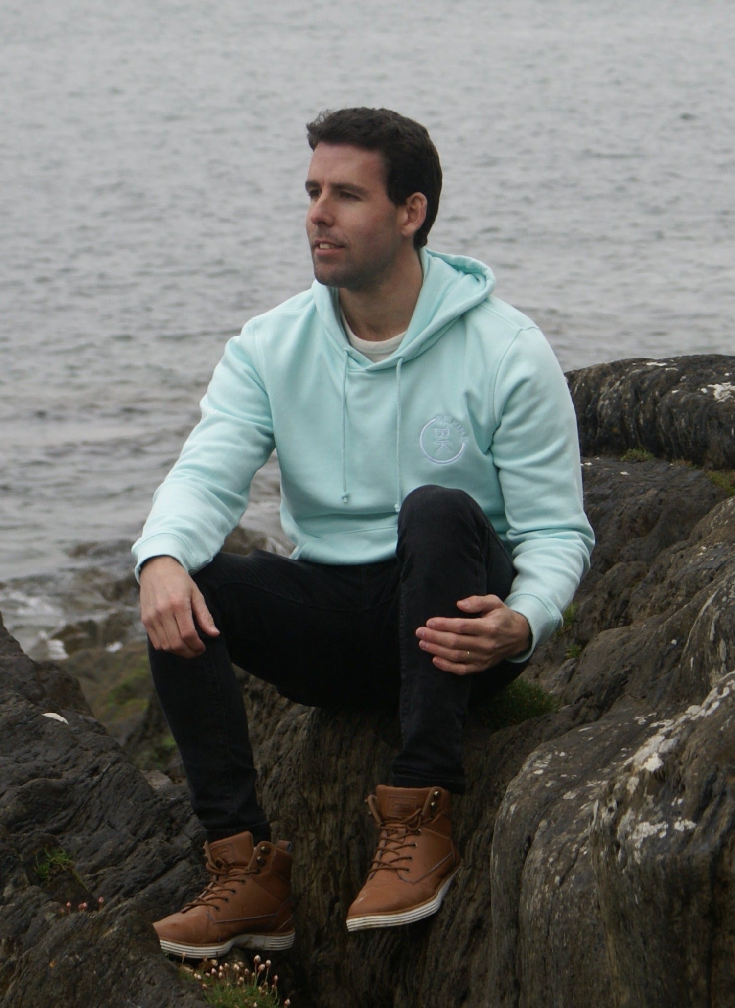 A man sits by the water on some rocks, he's wearing a Mint Green Organic Cotton Hoodie from the Be Kind Apparel Original Collection