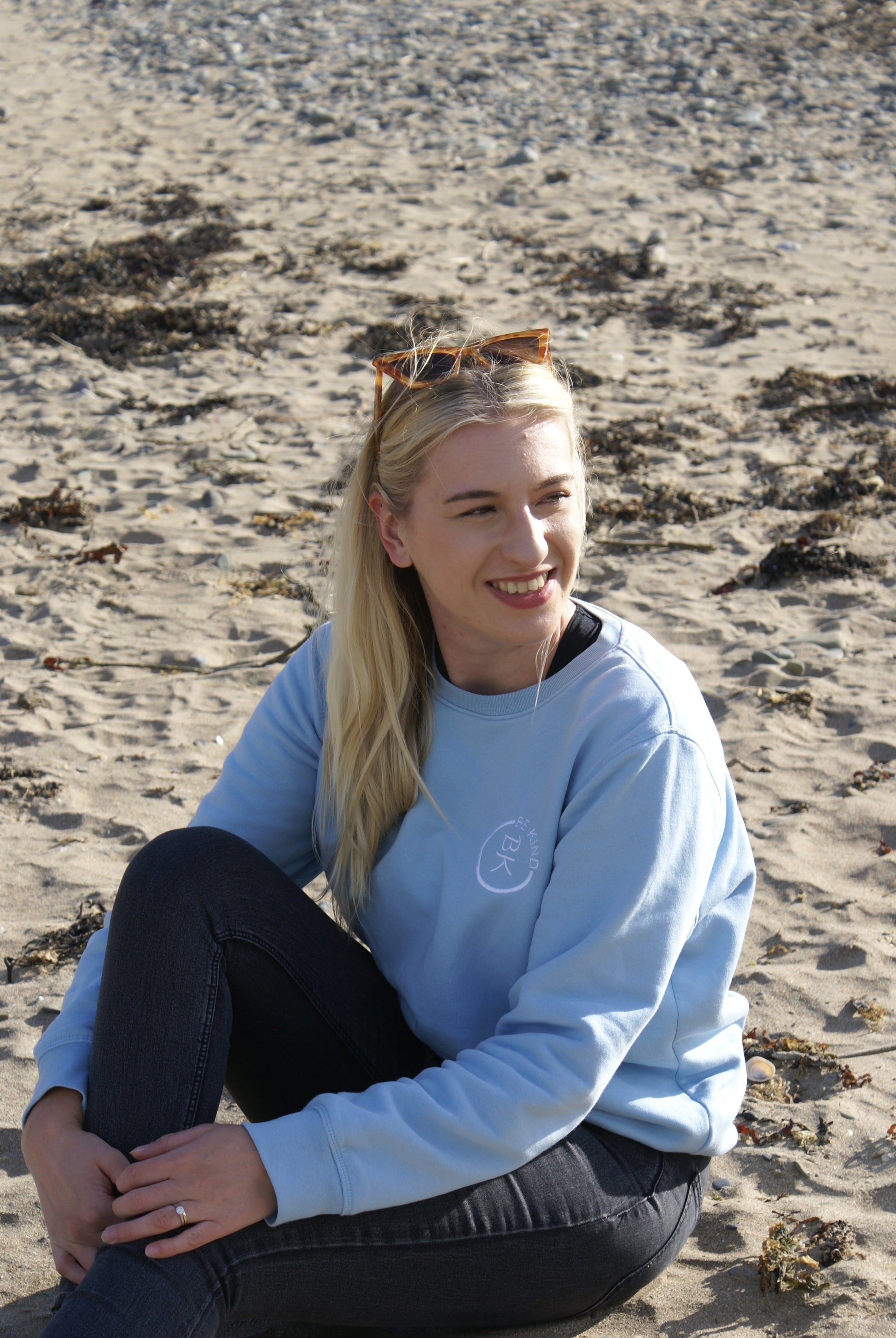 A woman sits on a beach, she's wearing a Sky Blue Organic Cotton Sweatshirt from the Be Kind Apparel Original Collection