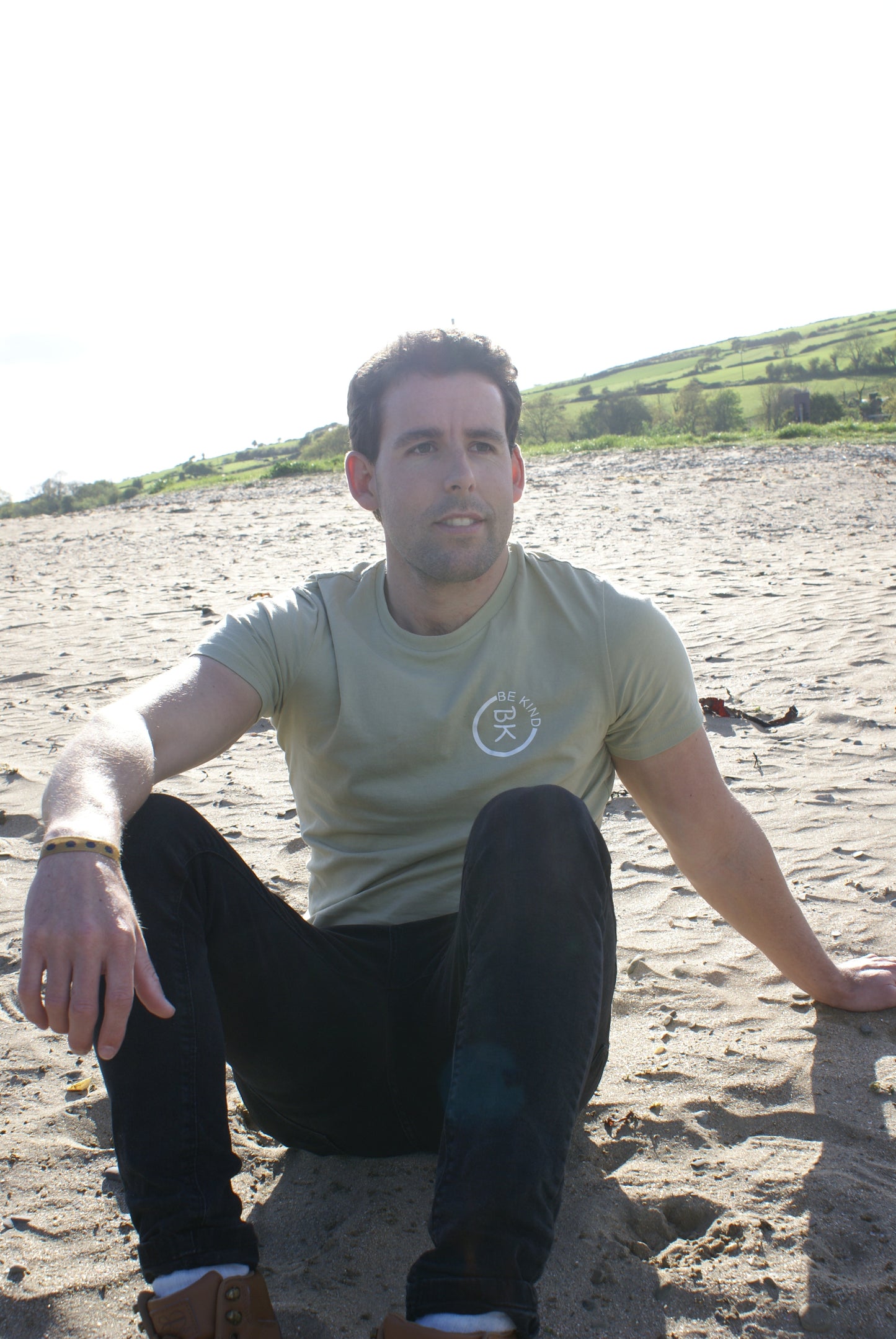 A man sits on a beach, he's wearing an Olive Green Organic Cotton T-Shirt from the Be Kind Apparel Original Collection