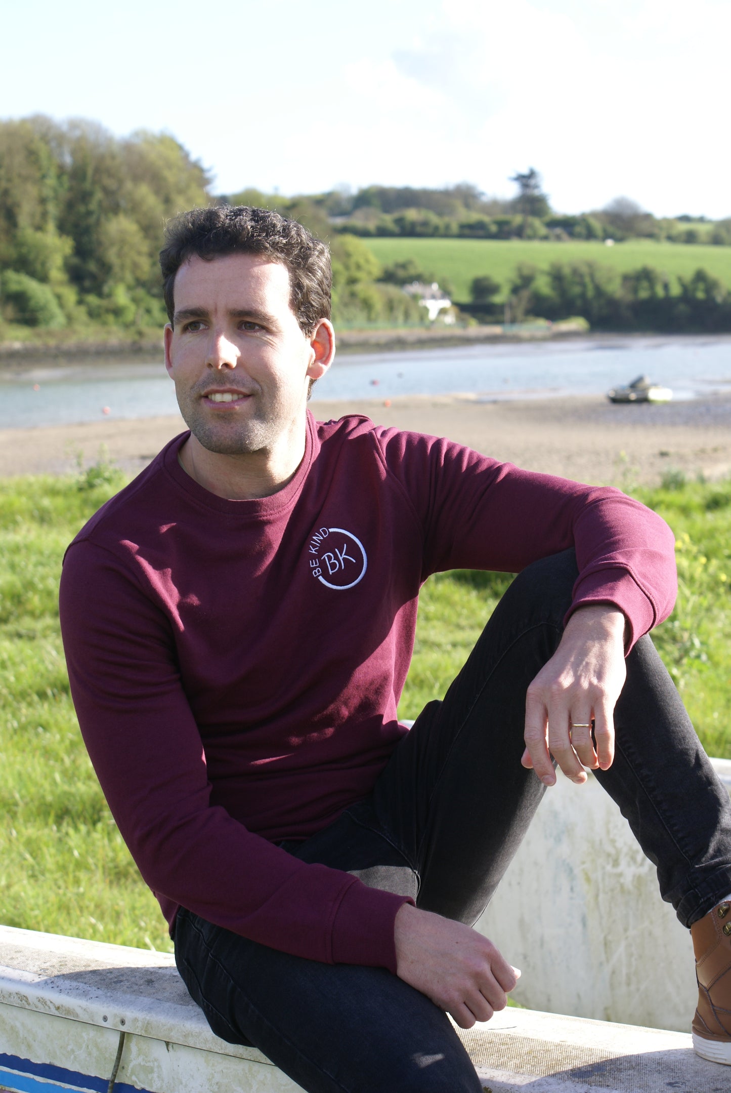 A man sits on the edge of a boat,  he's wearing an Organic Cotton Burgundy sweatshirt from Be Kind Apparel's Freestyle Range