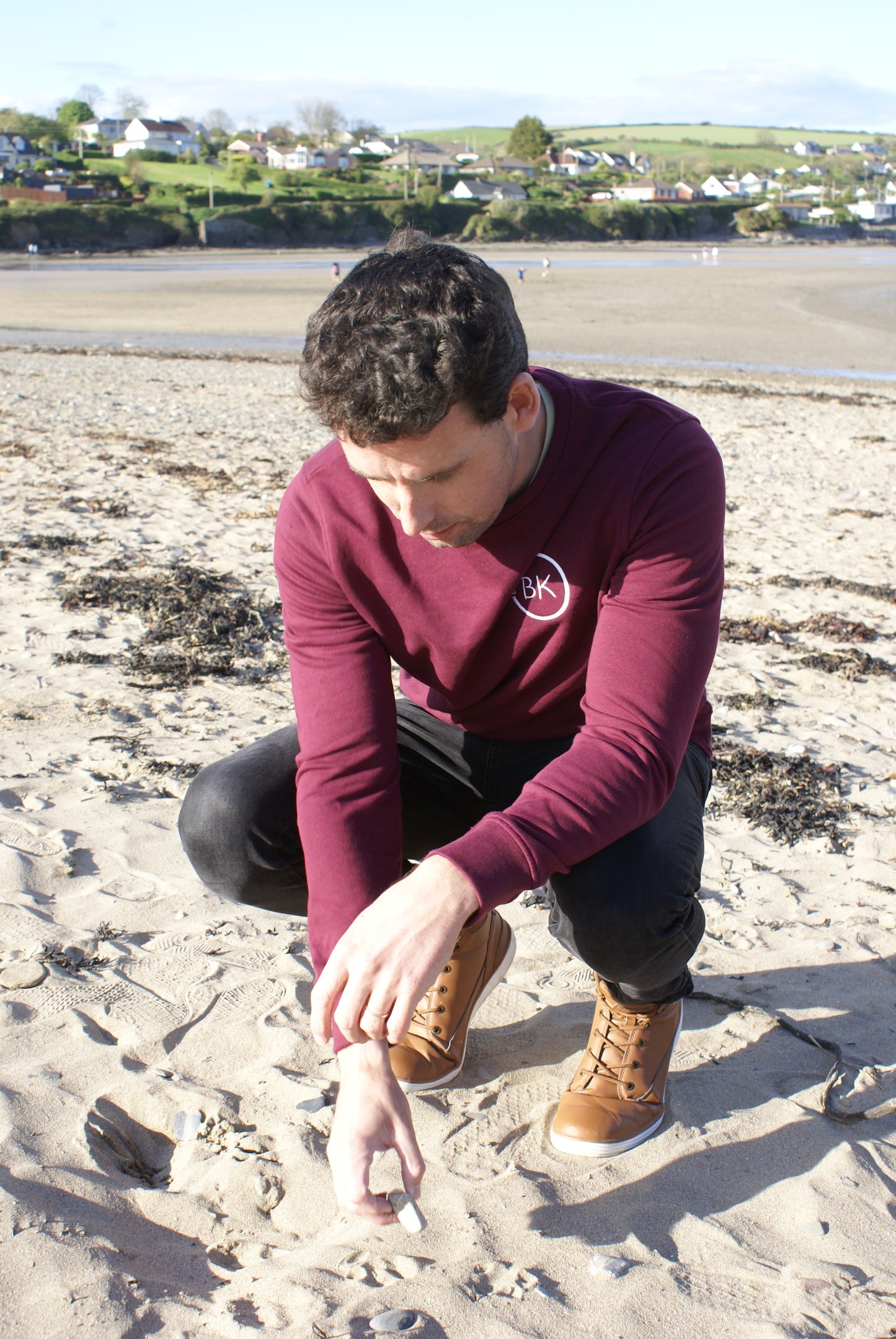 A man squats on a beach,  he's wearing an Organic Cotton Burgundy sweatshirt from Be Kind Apparel's Freestyle Range