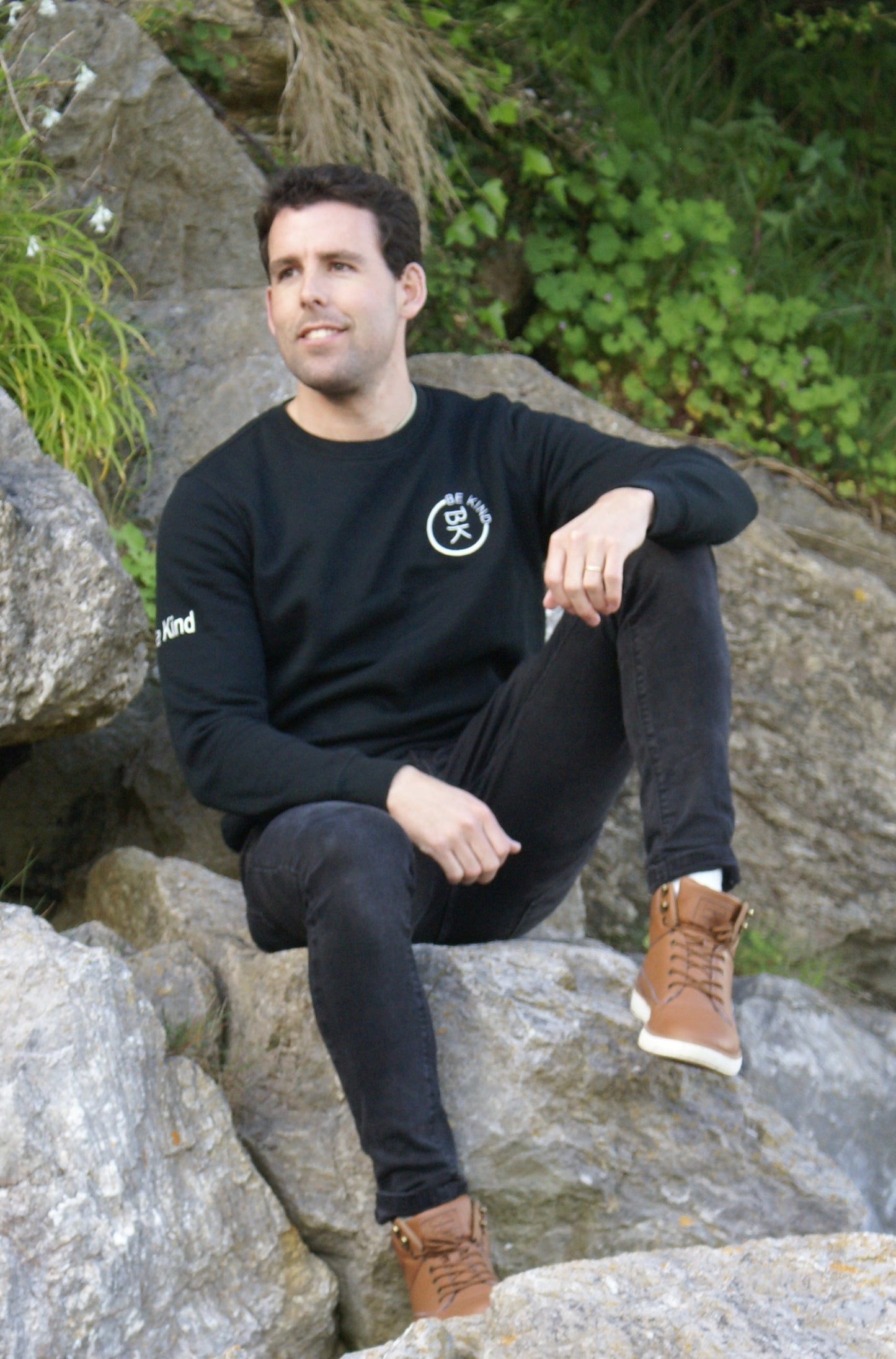 A man sits on some rocks, he's wearing a Black Organic Cotton Sweatshirt from the Be Kind Apparel Original Collection
