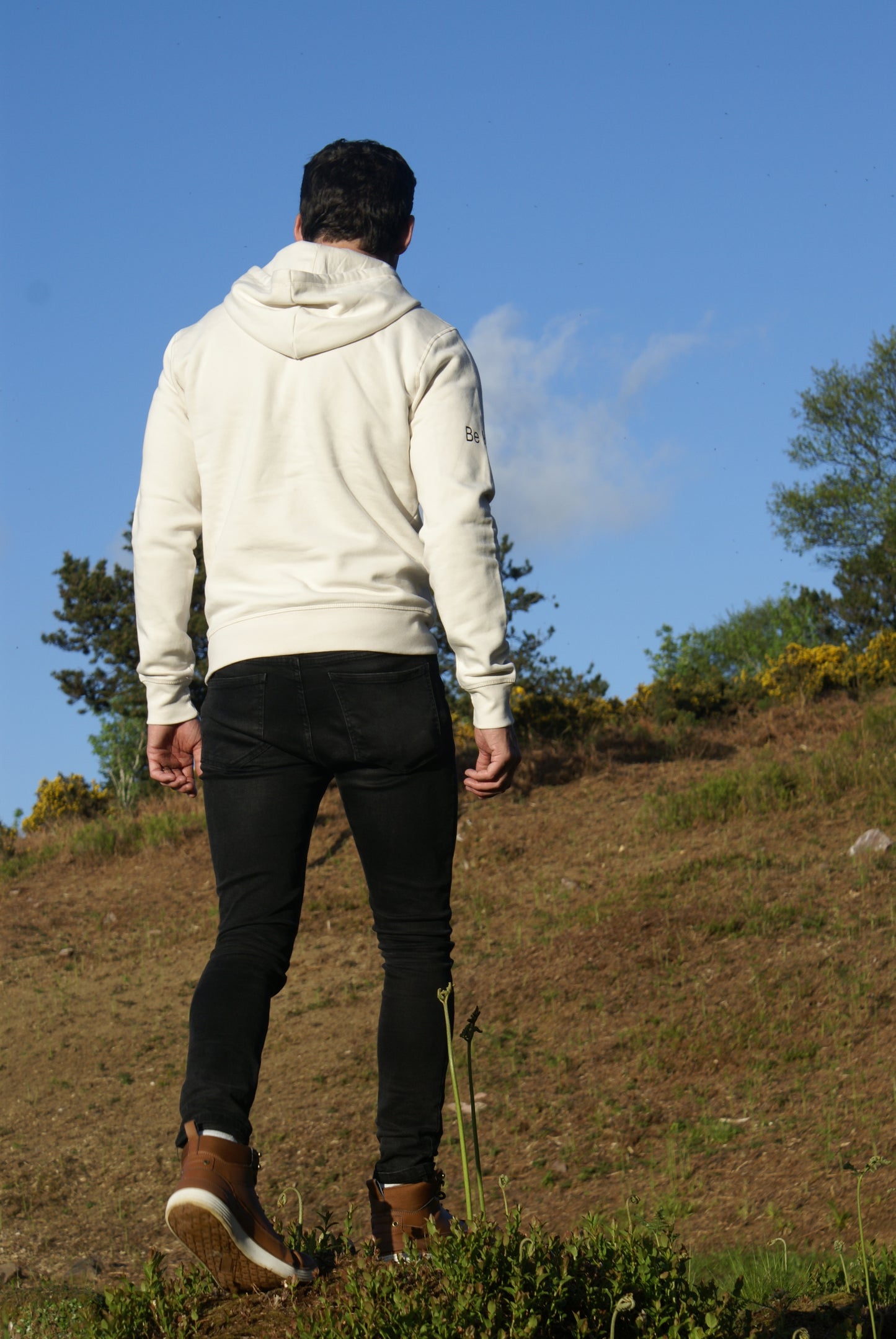 A man walks up a dirt hill, he's wearing an Off White Organic Cotton Hoodie from the Be Kind Apparel Original Collection