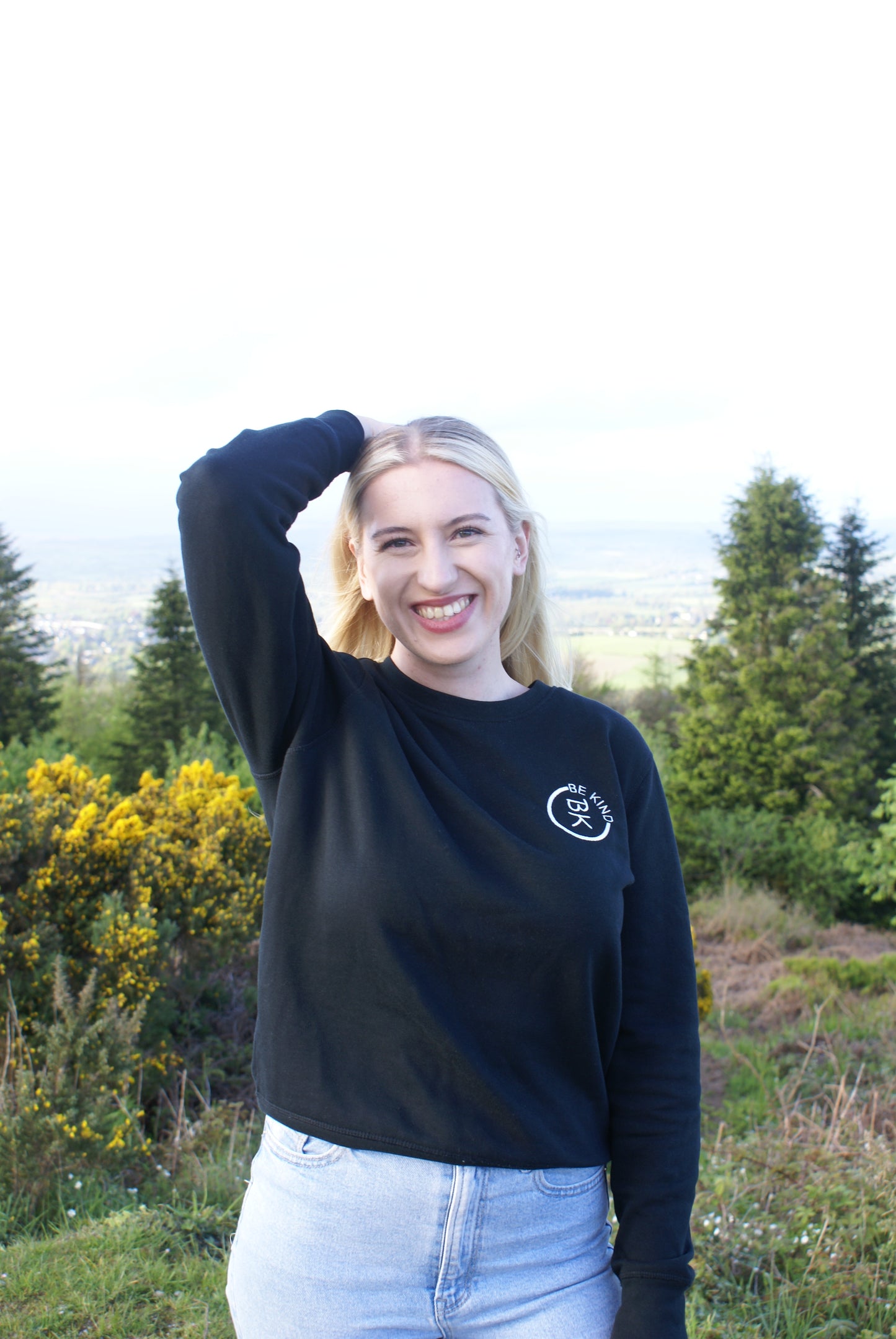 A woman smiles to the camera, she's wearing a Black Organic Cotton Sweatshirt from the Be Kind Apparel Original Collection
