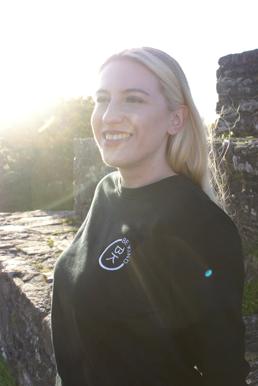 A woman smiles on a hill, she's wearing a Black Organic Cotton Sweatshirt from the Be Kind Apparel Original Collection