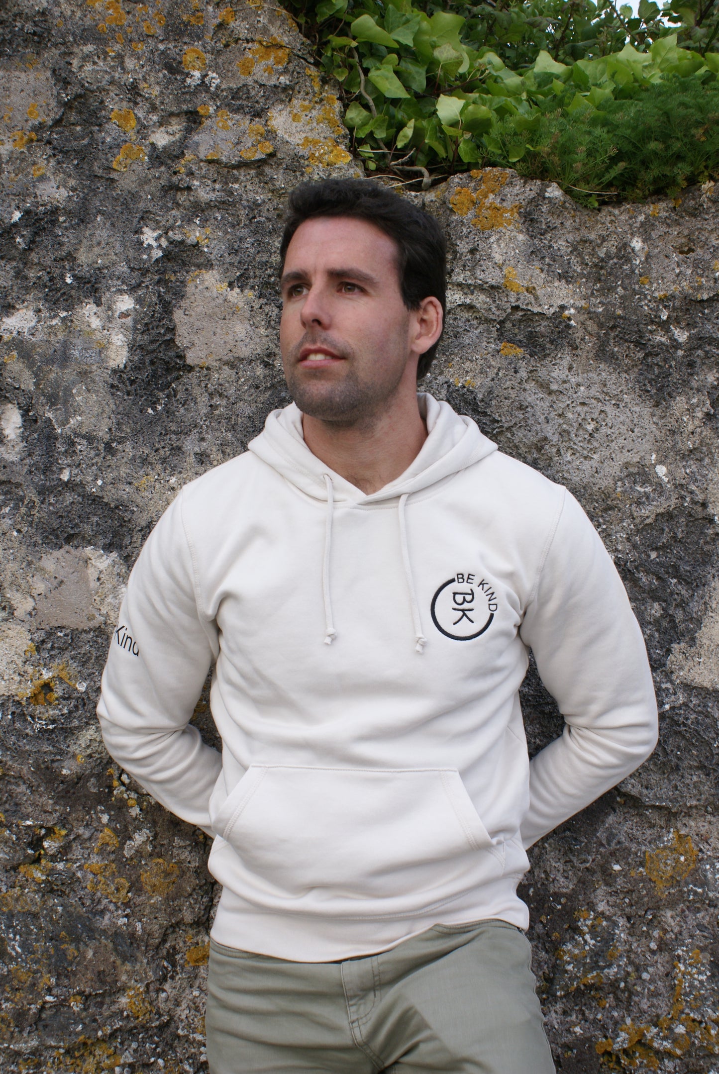 A man leans against a wall, he's wearing an Off White Organic Cotton Hoodie from the Be Kind Apparel Original Collection