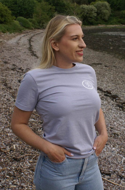 A woman stands on a beach, she's wearing a Lavender Organic Cotton T-Shirt from the Be Kind Apparel Original Collection