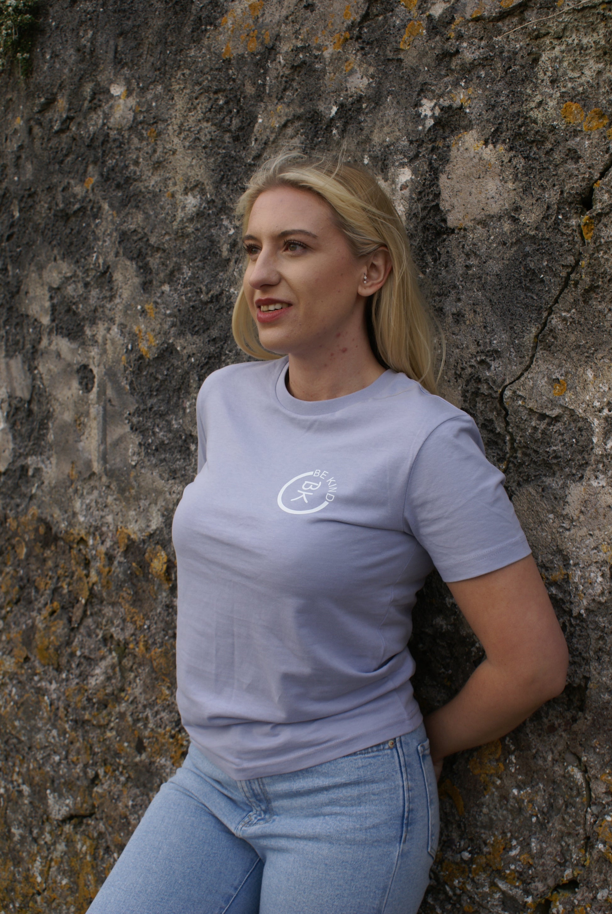A woman leans against a rock wall, she's wearing a Lavender Organic Cotton T-Shirt from the Be Kind Apparel Original Collection